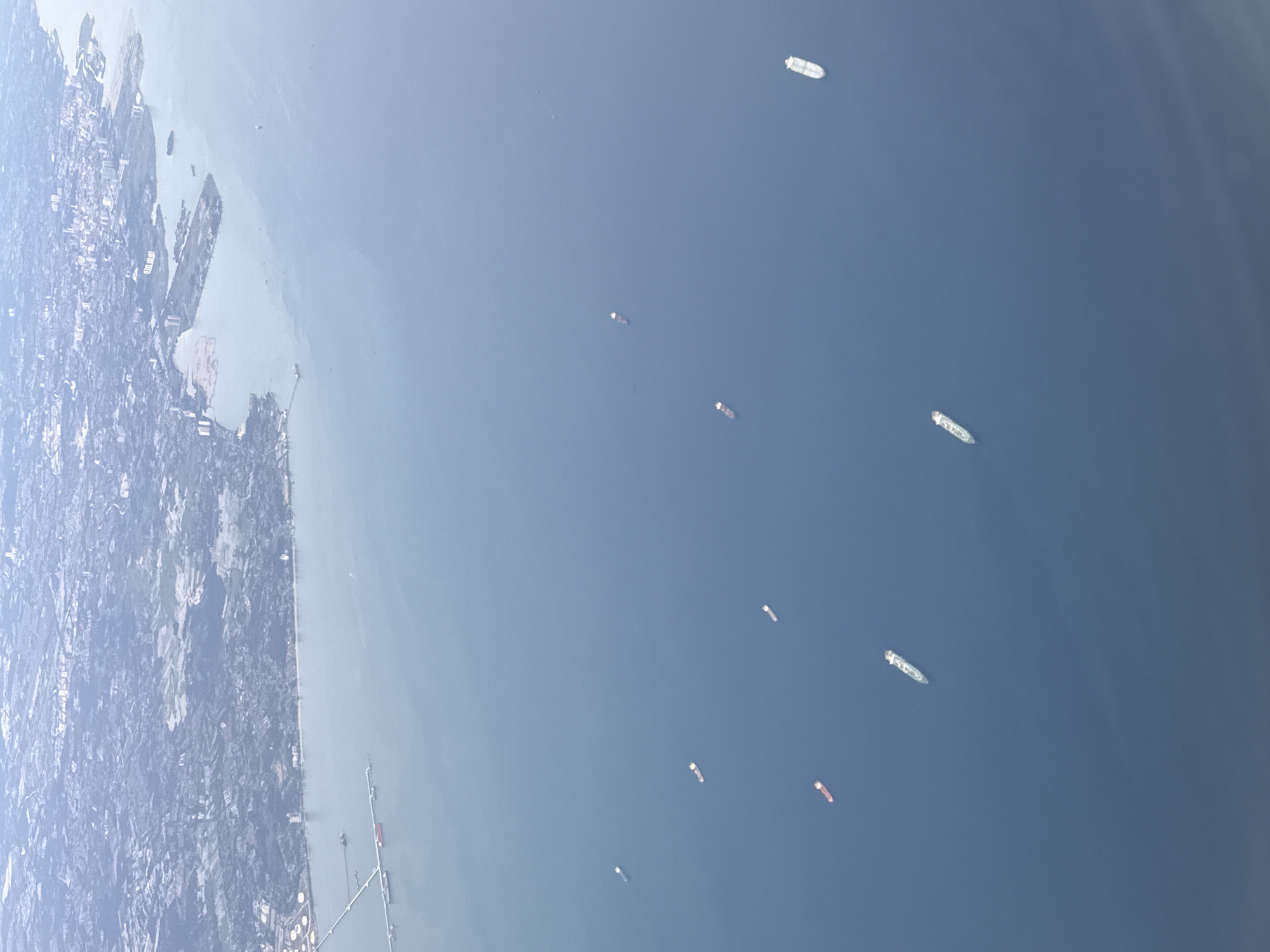 The image shows an aerial view of Denpasar, Indonesia. The coastline is visible on the left side of the frame, transitioning from a cityscape to a harbor with several piers. Numerous cargo ships are scattered across the ocean. The overall impression is one of a busy maritime environment with dense urban development along the coast.