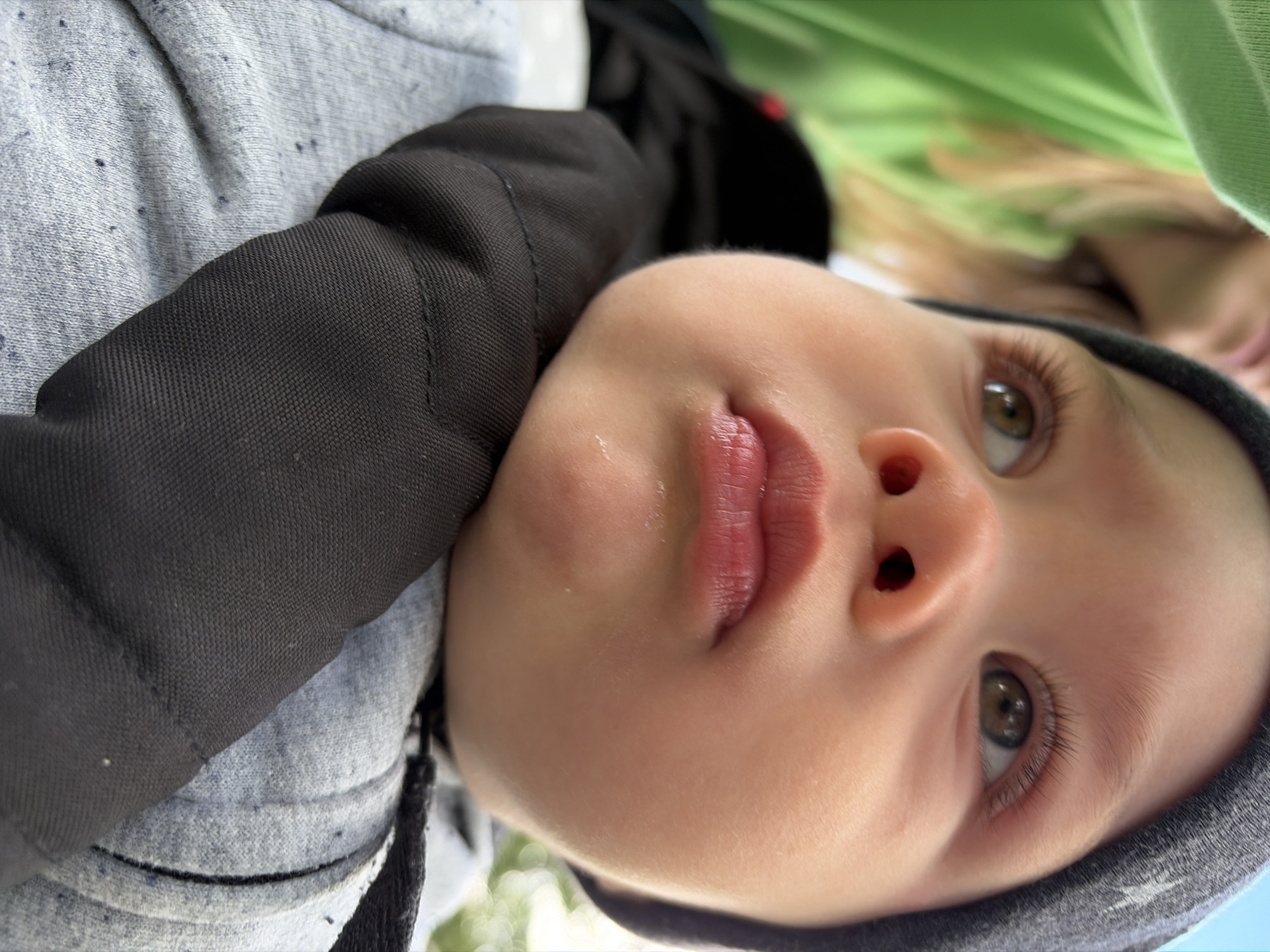 A close-up, rotated portrait-style shot captures a baby looking upwards with a serene and curious expression. The baby's face is central, showcasing prominent brown eyes with long eyelashes, a small nose, and full, slightly parted lips, with a tiny glistening droplet near the upper lip.
The baby is dressed in a light grey, speckled long-sleeved top, partially covered by a dark black garment, possibly a jacket or carrier strap, over their left shoulder. A dark grey hood or hat frames the baby's head.
In the highly blurred background, an adult or older child with light-colored hair is partially visible, wearing a bright green garment. Further in the background, out-of-focus green foliage can be seen, suggesting an outdoor setting or proximity to nature. The lighting is bright and natural, indicating daytime. The scene evokes a calm and tender moment. No text is visible. The location context is Chaikovsky, Russia.