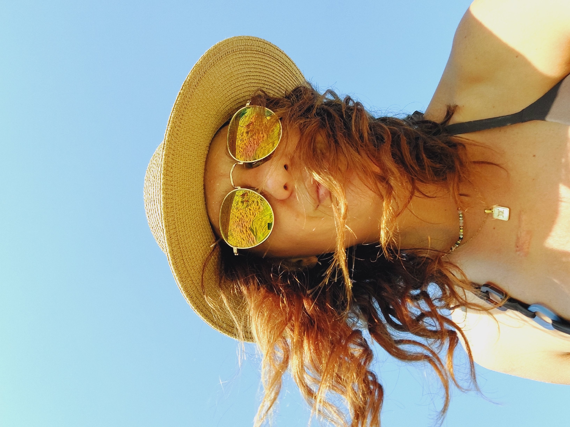 A low-angle, outdoor, waist-up portrait shows a woman wearing a straw hat and mirrored sunglasses against a clear blue sky. Her long, auburn hair is blowing to her left. The sunglasses reflect a beach scene with sand and water. She is wearing a necklace with a small, square pendant and a bikini top. The image was taken in Sveti Vlas, Bulgaria.