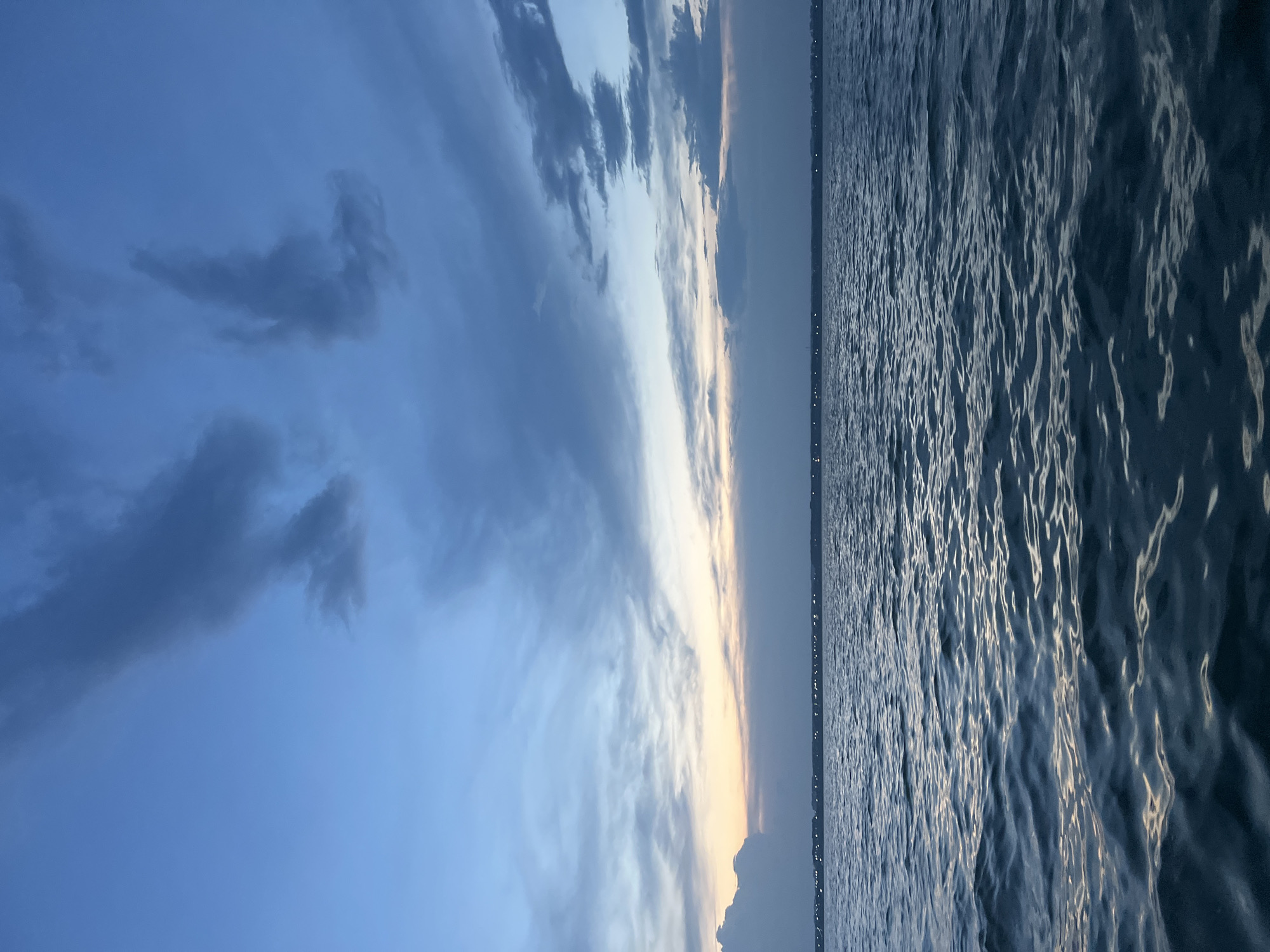 The image depicts a body of water, likely a river or a bay, under a twilight sky. The sky is a gradient of blues and grays, with scattered clouds that are dark and wispy. A faint band of pale orange and yellow is visible near the horizon, suggesting the sun has recently set or is about to rise. In the distance, a low-lying landmass is visible, dotted with numerous small lights that appear to be from buildings or streetlights, suggesting a settlement. The water's surface is rippled, with reflections of the sky creating shimmering patterns of light and dark. The overall impression is one of a tranquil, natural landscape transitioning between day and night. The location is identified as Novopichugovo, Russia. There are no people or specific objects clearly discernible in the foreground or midground.