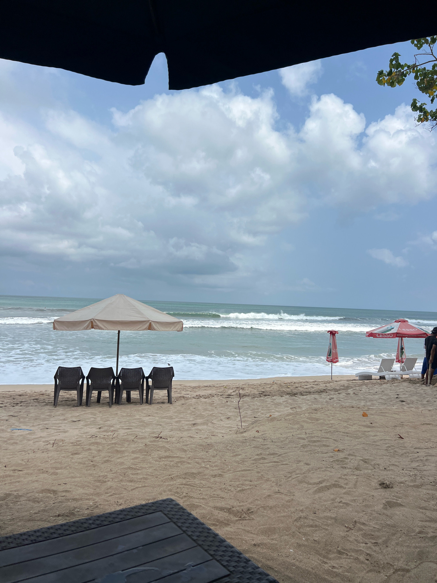 The image depicts a beach scene in Kuta, Indonesia, under a cloudy sky. In the foreground, a dark, woven table is partially visible. The middle ground features a sandy beach with several dark plastic chairs lined up under a light-colored umbrella. Further back, the ocean waves are breaking on the shore. To the right, two white lounge chairs are placed under red umbrellas. A few people are visible in the far right of the image, standing near the lounge chairs. The overall environment is a typical beach setting, with elements suggesting a relaxed atmosphere.