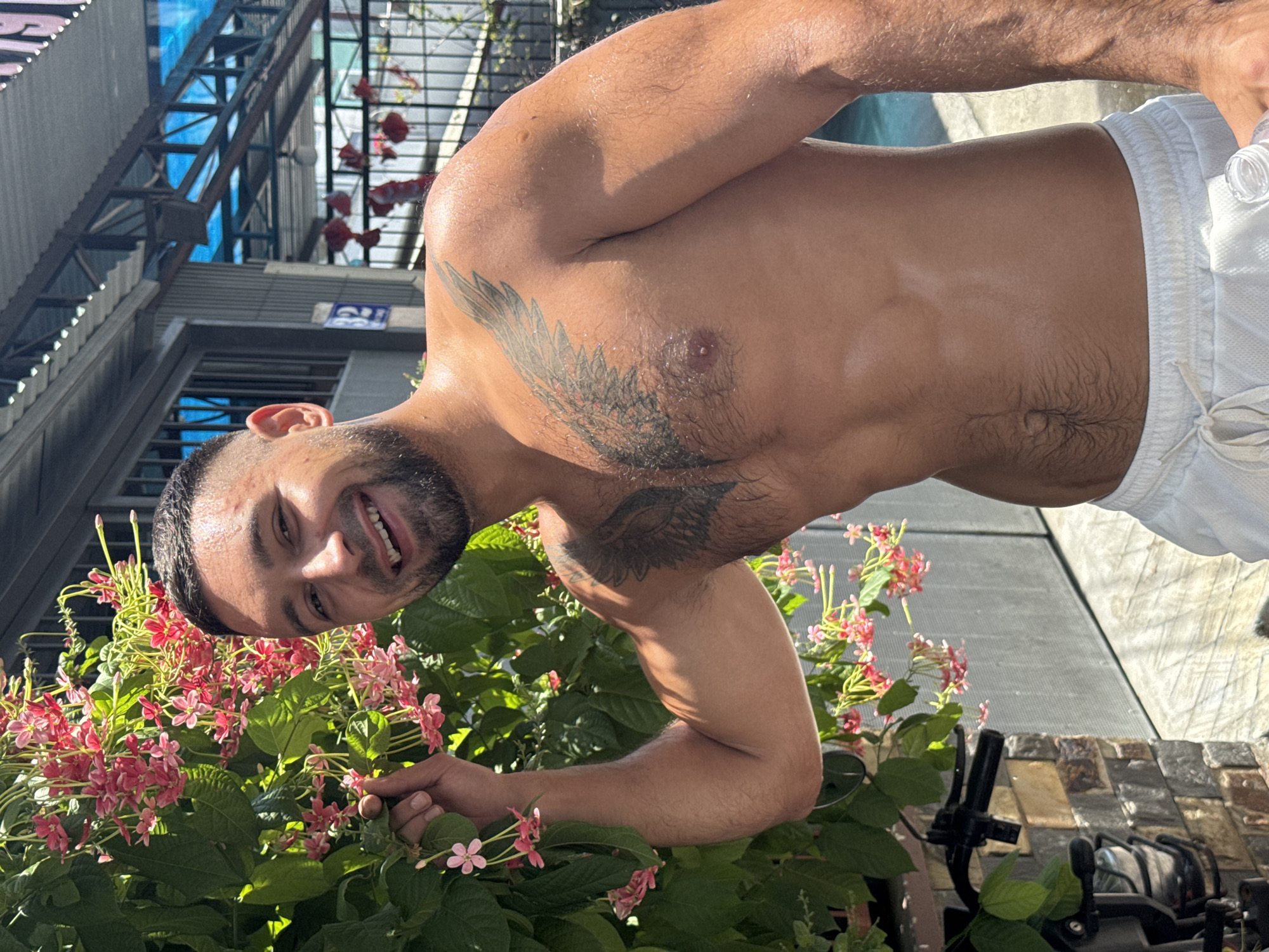 A shirtless man with a beard and a wing tattoo on his chest is standing in front of a flowering plant. He is smiling and holding a flower. The man is wearing white shorts. In the background, there is a building with a metal roof and a fence. A portion of a motorcycle is visible on the left side of the image. The setting appears to be outdoors, possibly in a residential area or a courtyard, with the environment suggesting a tropical climate, consistent with Nha Trang Ward, Vietnam.