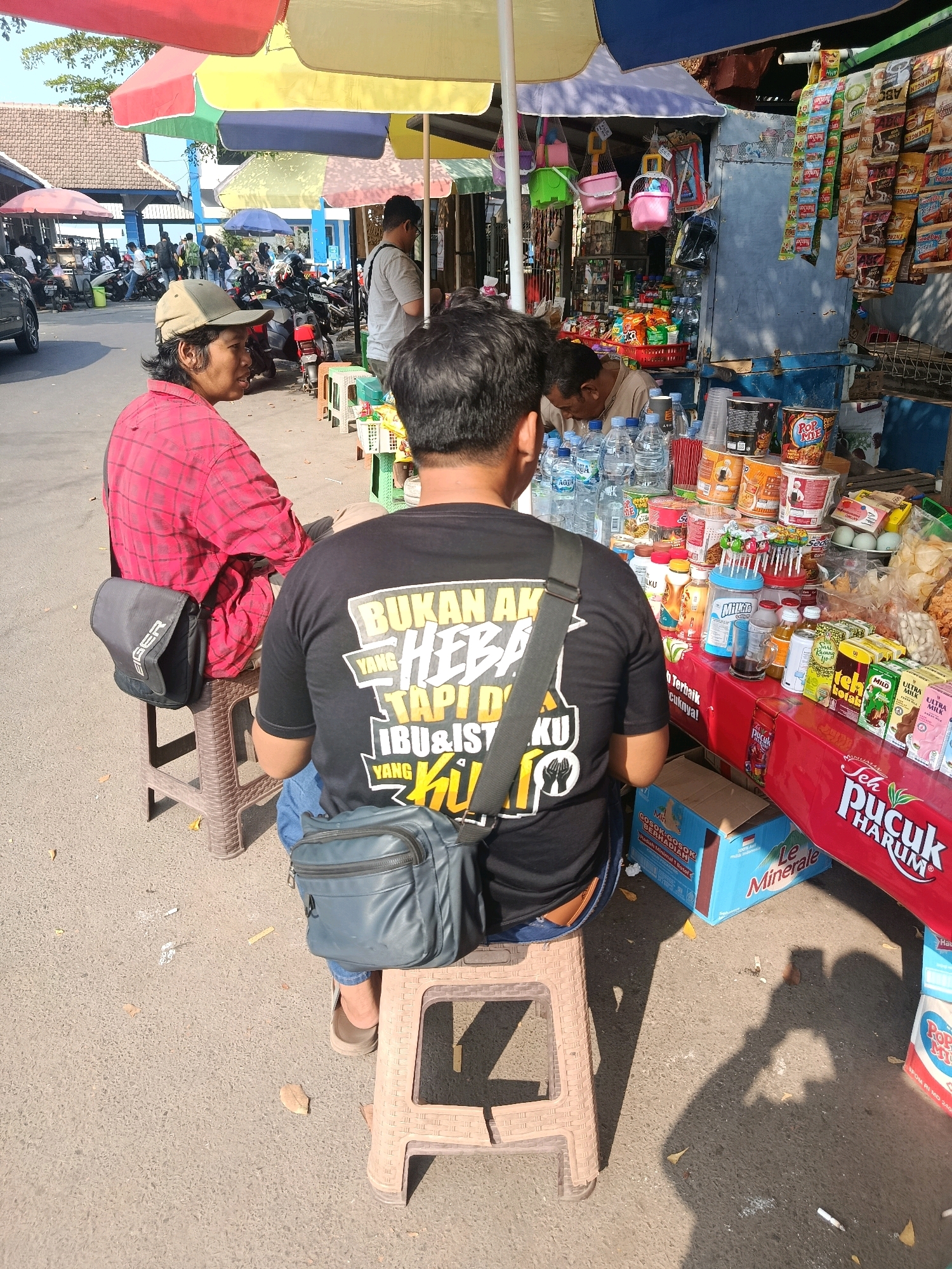 The image depicts a street scene in Jepara, Indonesia, focusing on a small roadside stall. Two individuals are seated on plastic stools in the foreground. The person on the left, wearing a red checkered shirt and a beige cap, has a dark grey sling bag on their left side. The person on the right, facing away from the camera, is wearing a black t-shirt with white and yellow text that reads "BUKAN YANG HEBAT TAPI IBUL IST YANG KUL." They also have a dark grey sling bag.
The stall behind them is laden with various goods, including bottled water, instant noodles, snacks, and packaged drinks. Multiple colorful umbrellas provide shade over the stall. In the background, there are other stalls, parked motorcycles, and a few other people, suggesting a bustling marketplace or roadside commercial area. The overall environment appears to be outdoors, with sunlight casting shadows on the ground.