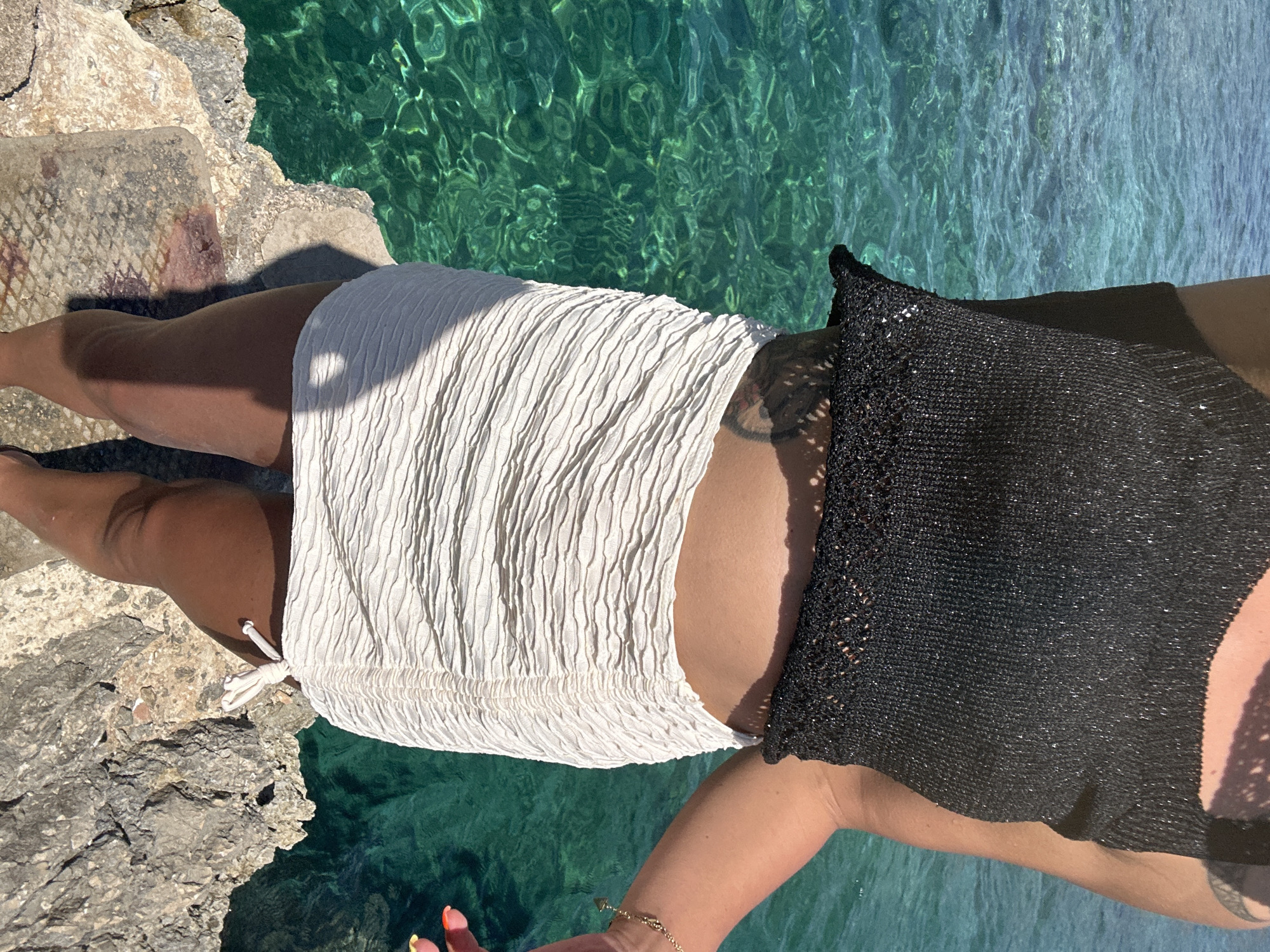 The image shows a person standing on rocks near the water in Cala Lliteres, Spain. The person is wearing a black metallic knit crop top and a white ruched mini skirt. The sea behind them is a vibrant turquoise color. The sun is shining, casting shadows on the rocks and the person's legs. The person has a tattoo on their hip and is wearing a gold bracelet. Their fingernails are painted with neon colors. The rocks are grey and textured, with some patches of red earth.
