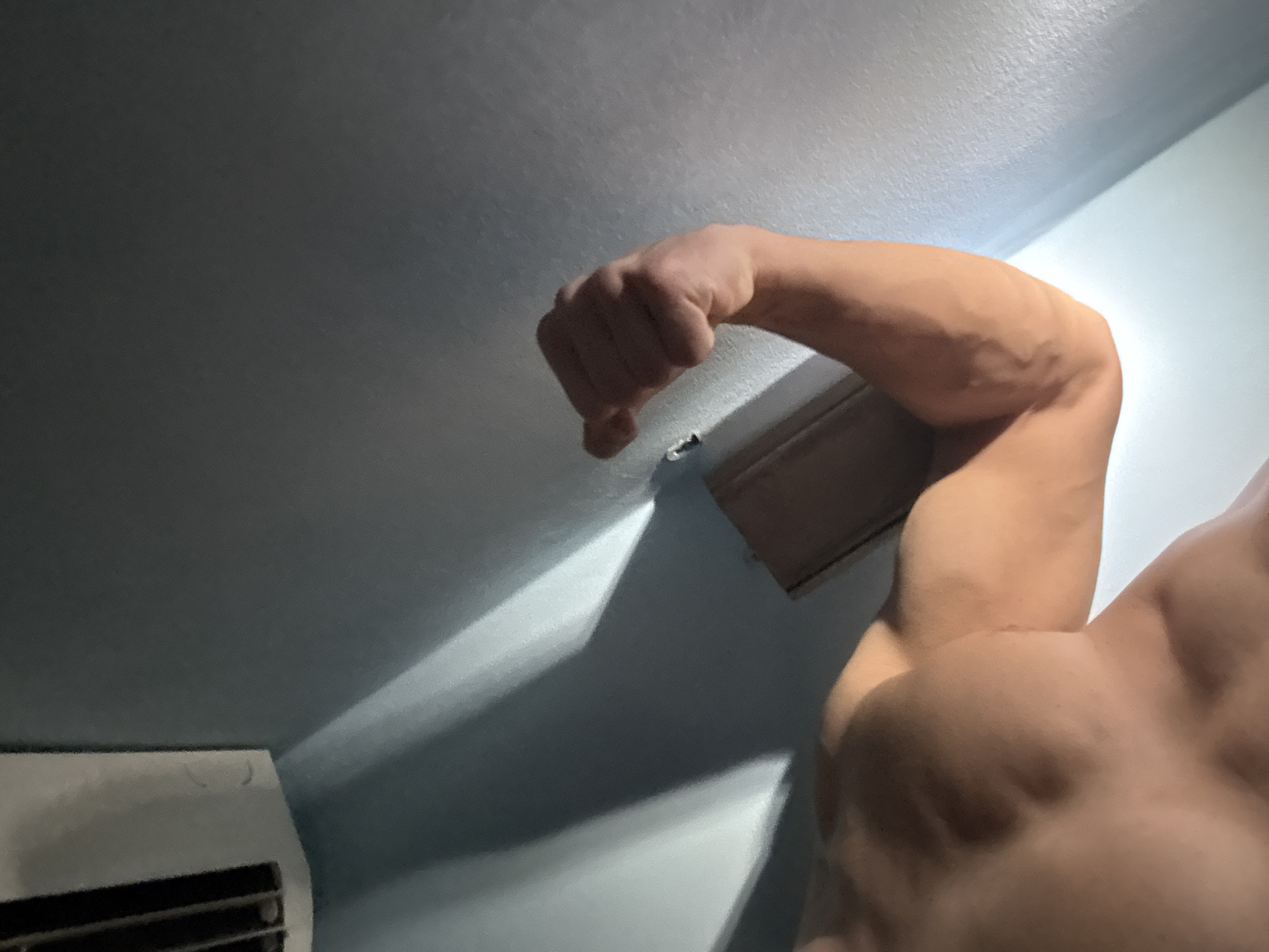 A low-angle, close-up shot of a flexed arm and chest. The fist is clenched and raised towards a light-colored ceiling with molding. The arm muscles are defined and the chest is partially visible in the lower right. The background is a pale blue wall with shadows. An air conditioning unit is visible in the lower left corner. No identifying features indicate the location as Buhovo, Bulgaria.