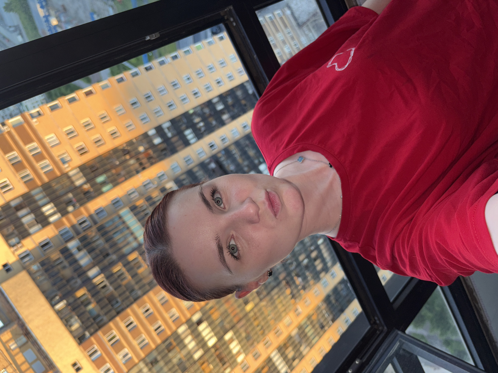 An adult female with auburn hair pulled back is taking a selfie. She is wearing a bright red t-shirt with a small white heart design on the left breast. She has green eyes and is puckering her lips. She is wearing small black earrings and a thin silver necklace with a blue quatrefoil pendant. The background consists of a blurred cityscape with tall, modern residential buildings, some illuminated by golden hour sunlight. The scene appears to be indoors, possibly on a high floor with a large window or glass enclosure. The overall impression is a casual, personal photograph taken from a high vantage point in a city.