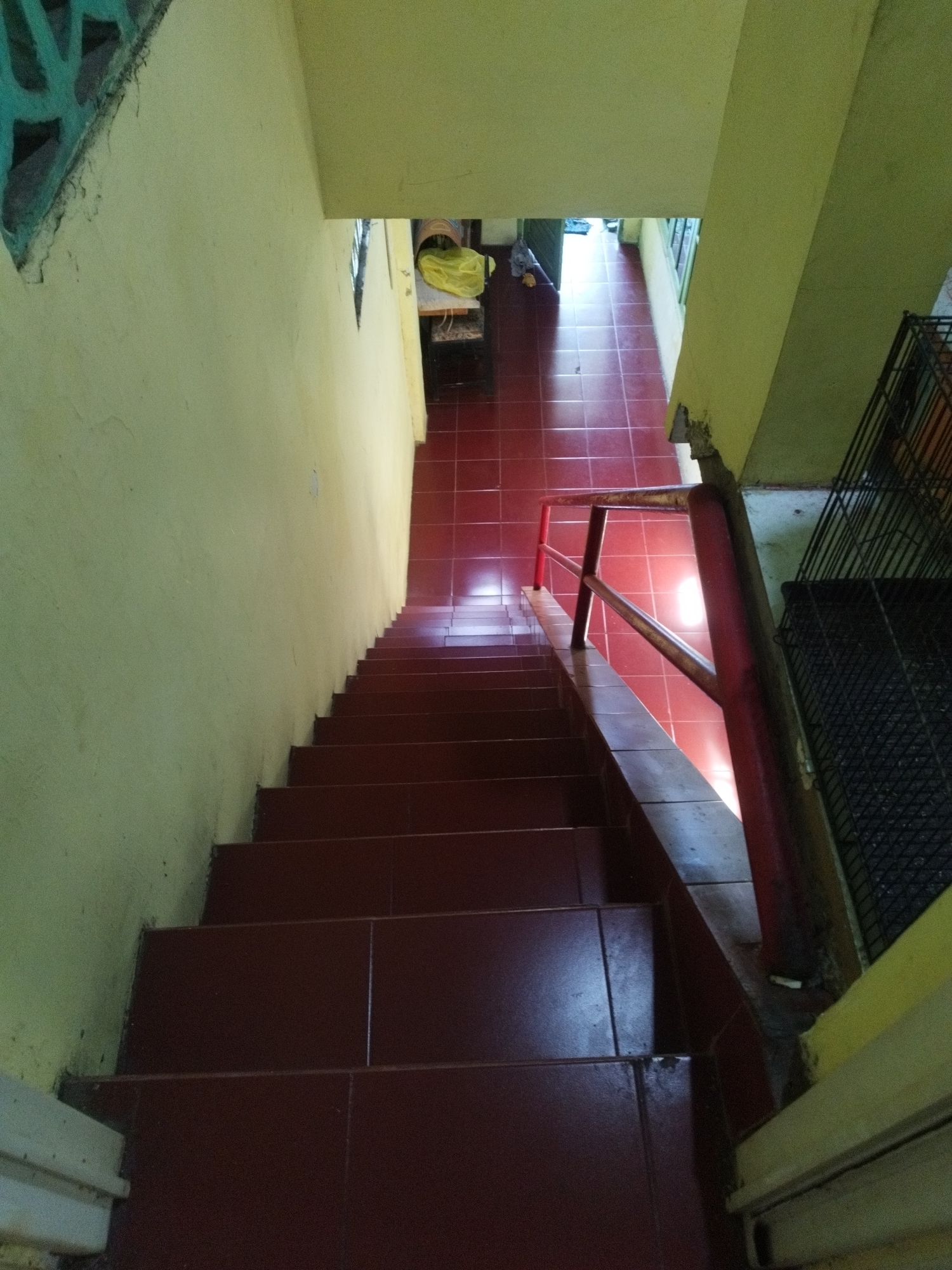 The image shows a dimly lit indoor staircase with maroon tiled steps and walls painted a pale yellow. A red metal handrail runs along the right side of the stairs. The staircase descends into a hallway with red tiled flooring, leading towards an open doorway that reveals daylight. To the right of the stairs, there is a black metal grate structure. The scene appears to be located in Sumenep, Indonesia, likely within a residential or commercial building, given the architectural style and materials.