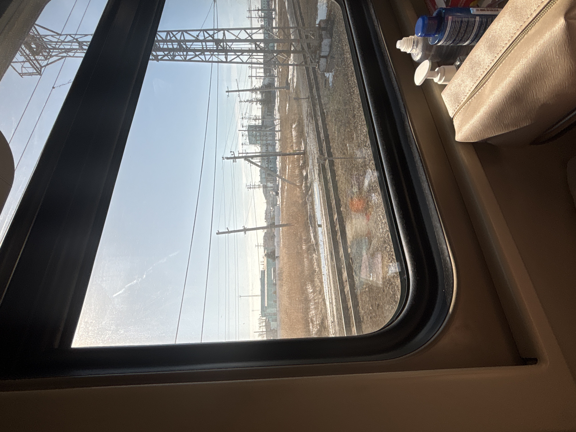 The image provides a view from inside a train, looking out a window onto a railway environment in the city of Kalachinsk, Russia. The scene is illuminated by bright daylight under a clear, light blue sky.
Inside the train, a section of a beige interior wall and the dark-framed window are visible. On the window's lower ledge, a light beige/peach-colored zippered pouch is placed, behind which stand three bottles: a white pump-dispenser bottle, a clear plastic bottle with a silver cap, and a blue plastic bottle with a blue cap. No individuals are present in the frame.
Outside the train, multiple parallel railway tracks extend across the mid-ground. The ground surrounding the tracks is covered with dormant, light brown vegetation and scattered patches of snow or ice, indicating a late winter or early spring setting. Numerous utility poles, some wooden and others metal, support a network of overhead wires that crisscross the sky. A tall, dark metal lattice tower, likely a transmission or communication structure, is prominent on the right side of the view. In the background, several industrial or commercial buildings with flat roofs are discernible, one of which features a distinct turquoise or light green facade. Further utility infrastructure and sparse, bare trees are also present in the distance.