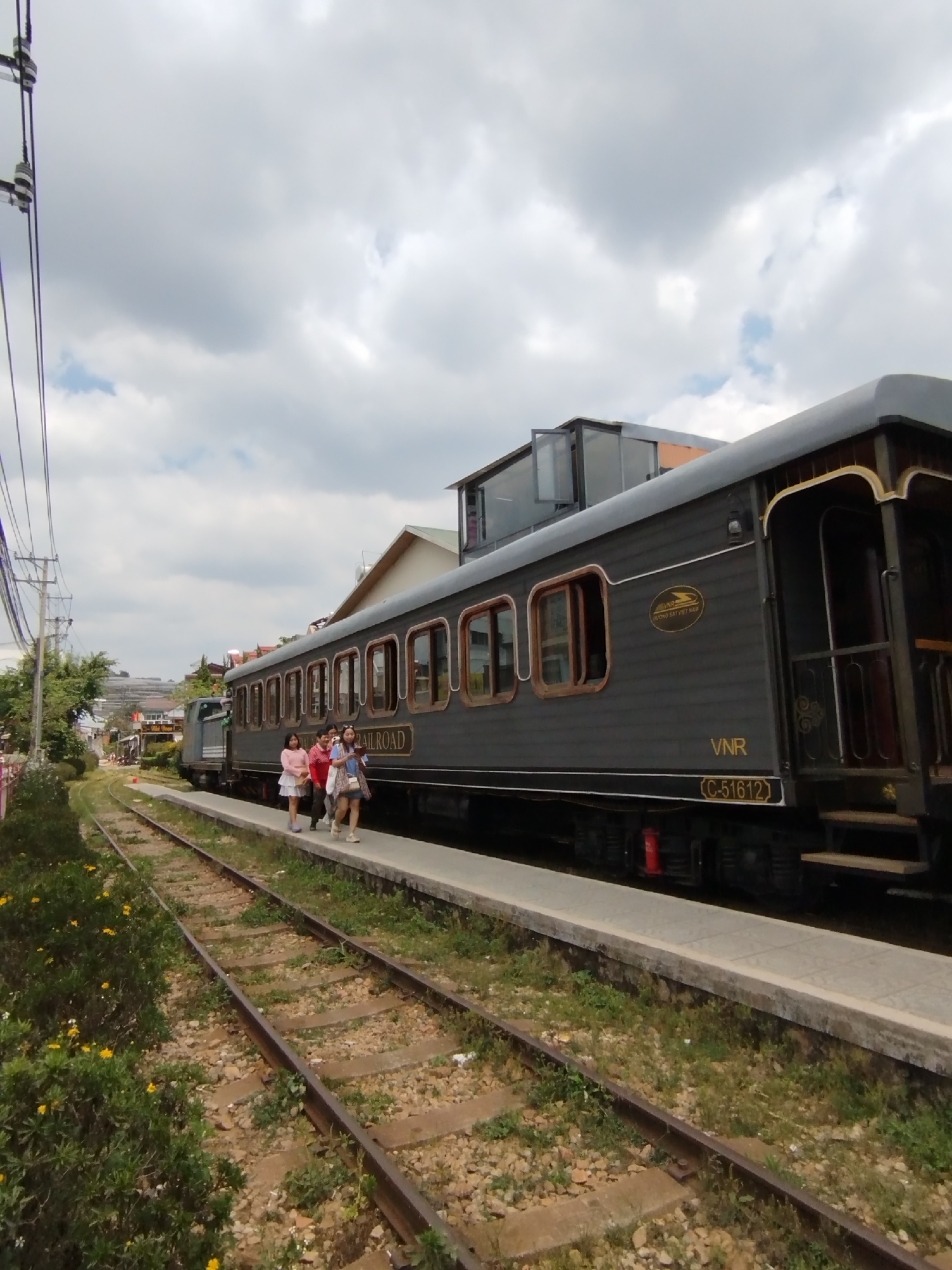 A railway car is parked on a platform next to a rail track. The car is painted a dark gray color with gold accents around the windows and the end entrance of the car. The side of the rail car reads "RAILROAD." Three people are walking along the platform next to the car. The platform is a light gray concrete and extends along the length of the train car. Bushes with small yellow flowers are growing next to the platform and the rail tracks. The railway is in Lộc Quý, Vietnam. Buildings are visible in the background, including a two-story building with a modern glass-enclosed roof. The sky is overcast with heavy clouds. Electrical wires are visible to the left of the frame.