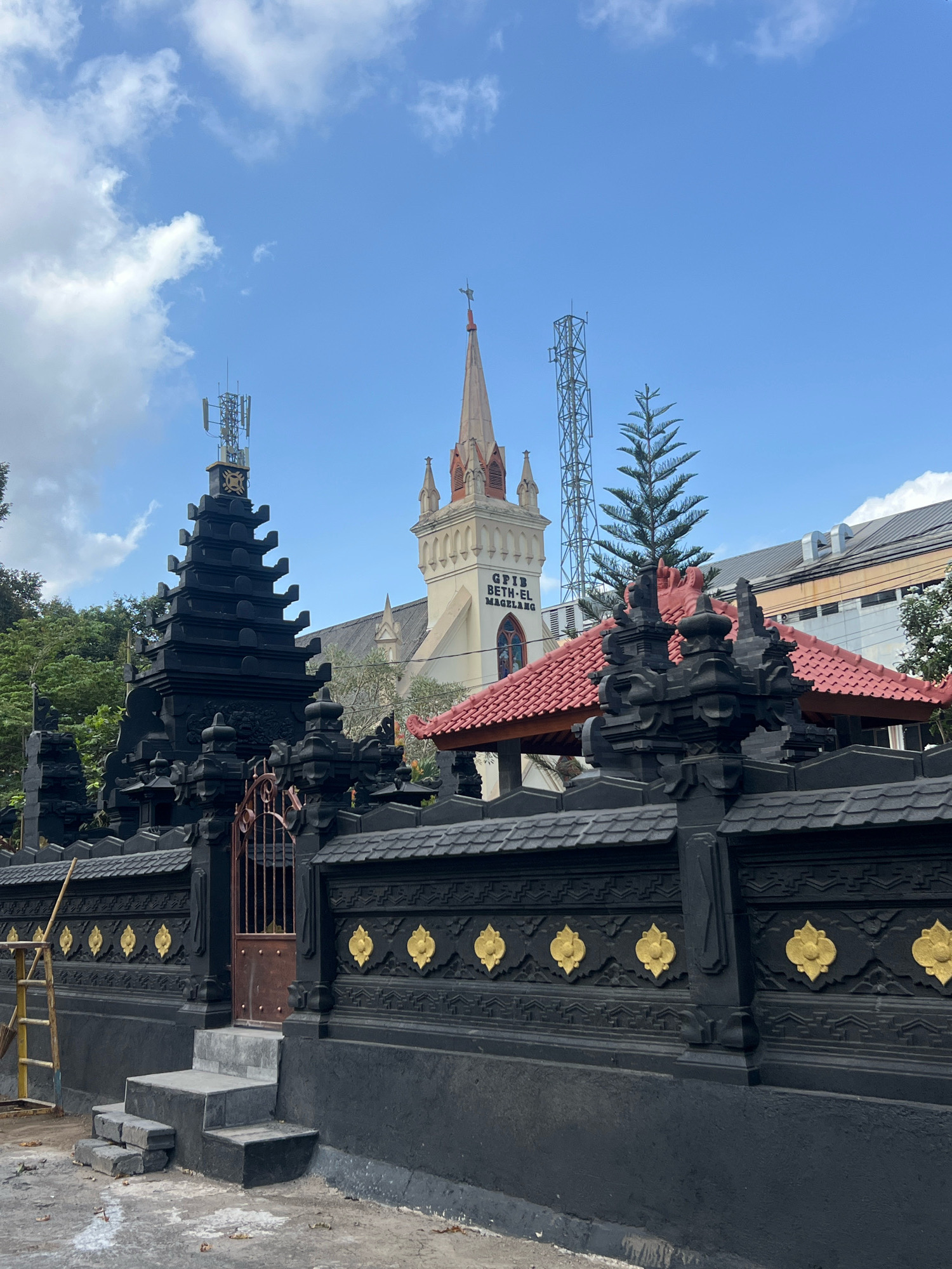 The image shows a view of GPIB Beth-El Magelang, a church in Magelang, Indonesia, with a striking black stone fence in the foreground. The fence features intricate carvings and ornate gold floral embellishments. A rustic metal gate is set within the fence, with a set of stone steps leading up to it. Beyond the fence, the church's facade is visible, characterized by a tall spire topped with a cross and a section of text reading "GPIB BETH-EL MAGELANG". A red-tiled roof structure is also present. The sky above is blue with scattered white clouds, and a tall, slender pine tree stands near the church. Several communication towers are also visible in the background, suggesting an urban or semi-urban setting. Green trees surround the church building.
