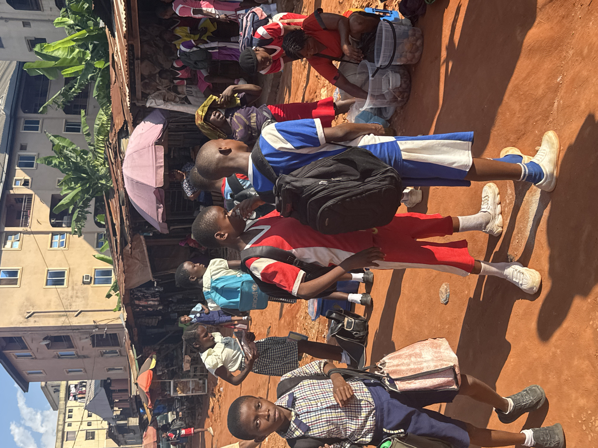 The image depicts a group of children, likely students, gathered in an outdoor market setting. The children are dressed in various school uniforms, some in red and white, others in blue and white, and one in a plaid skirt and light-colored top. They appear to be waiting or milling about, with some carrying backpacks.
In the background, a market stall is visible with shelves stocked with goods, and a pink umbrella providing shade. Behind the market, multi-story buildings with windows and balconies are seen, suggesting an urban environment. Banana trees are also present, indicating a tropical or subtropical climate.
The ground is a reddish-brown earth, and the scene is illuminated by bright sunlight, casting distinct shadows, suggesting it is daytime. The children's expressions are mostly neutral, with one child in the foreground looking directly at the camera. The overall impression is one of everyday life in a busy marketplace. There are no clearly visible text elements in the image.