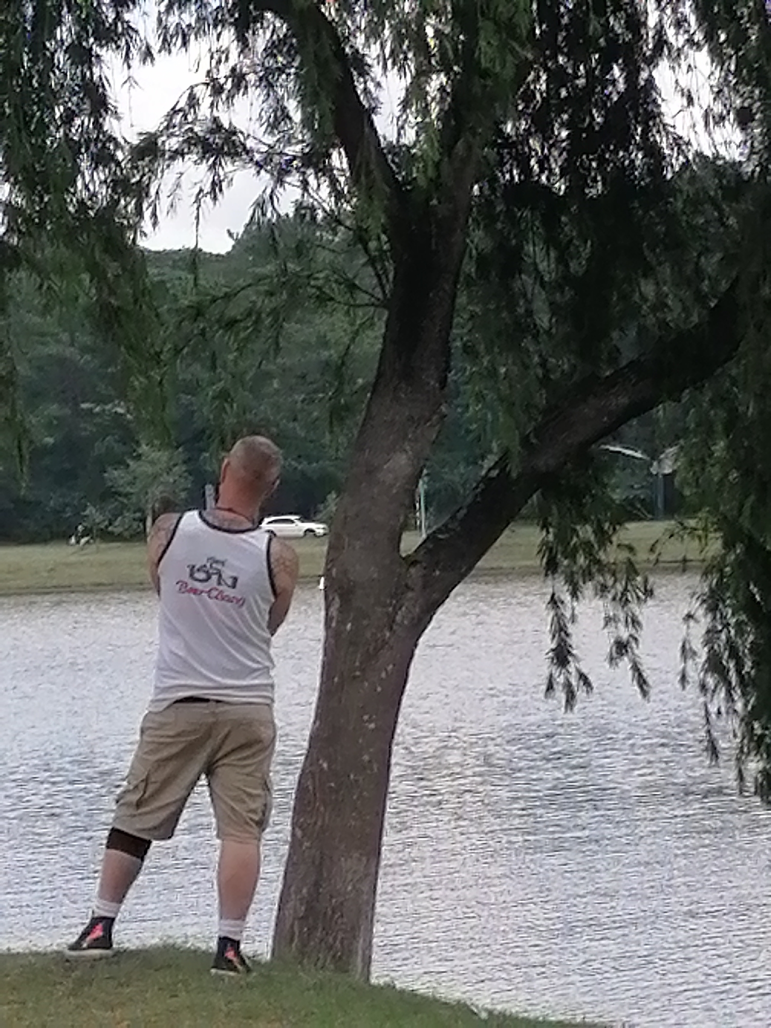 A person stands on a grassy bank facing a body of water, likely a lake or pond. The person is viewed from the back and is wearing a white tank top with a graphic design and text that appears to be in a foreign script, along with tan cargo shorts. They have tattoos on their arms. The person is wearing a knee brace on their left leg and athletic shoes.
The scene is dominated by a large tree trunk in the foreground, with its branches and foliage obscuring part of the sky and background. The sky appears overcast, suggesting a cloudy day. In the distance, across the water, there are trees and vegetation, forming a backdrop of greenery. A white car is visible parked on a road in the background. The water is calm, with ripples indicating a gentle breeze. The overall setting suggests an outdoor recreational area, possibly a park. There are no clear indicators of the time of day, but the lighting suggests it is daytime. The emotional tone of the image is neutral, focusing on the individual and the natural environment. Visible text on the person's tank top includes what appears to be Asian characters and "Beer-Classy".