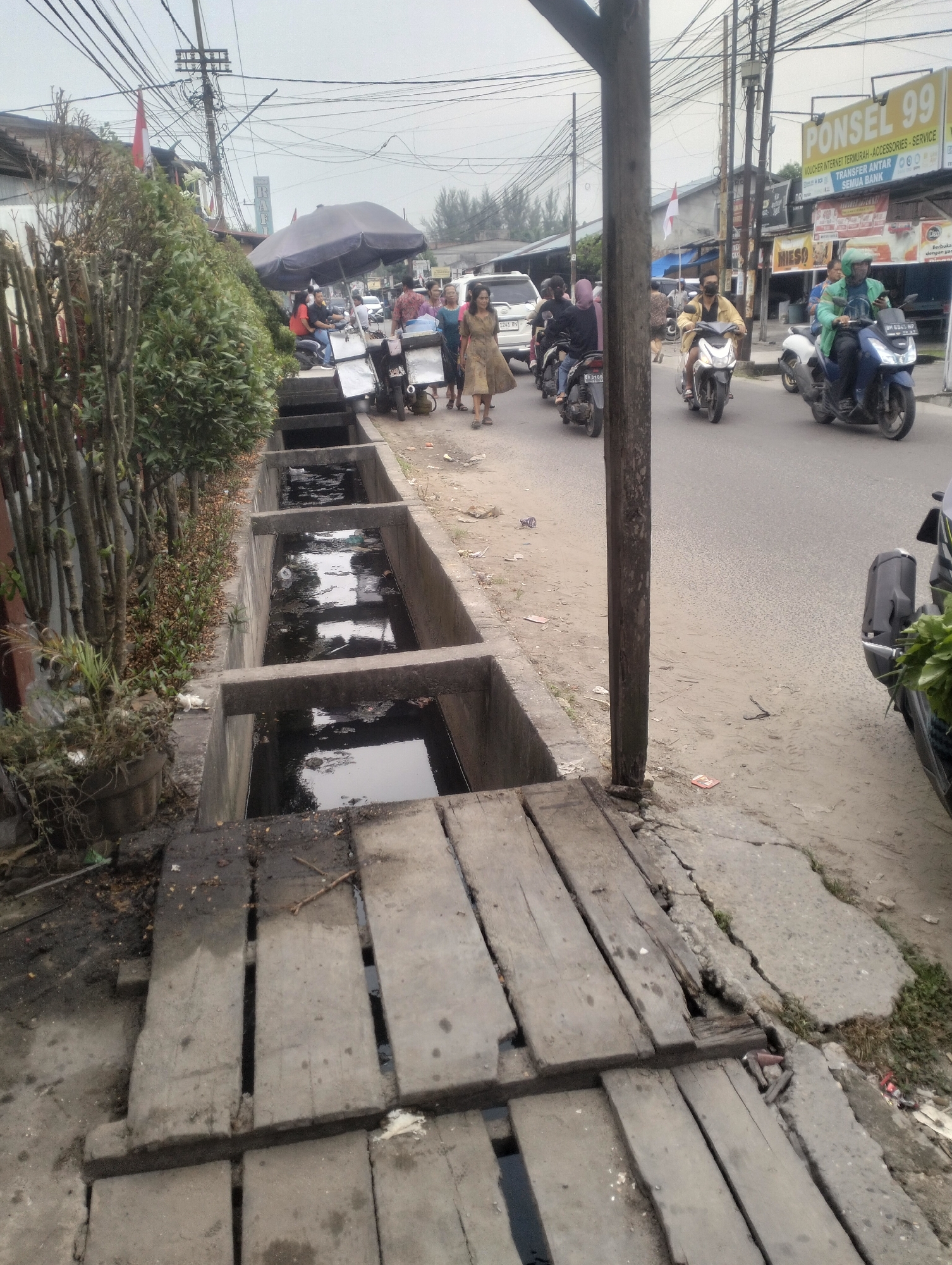 The image depicts a street scene in Dumai City, Indonesia. A concrete drainage ditch runs along the left side of the road, partially covered by wooden planks. The ditch appears to contain murky water and debris. The road itself is paved and shows signs of traffic. Several motorcycles are visible, some carrying passengers, on the road. A few pedestrians are also present, including a woman in a light-colored dress walking on the sidewalk. On the right side of the road, there are buildings with storefronts, including a sign for "PONSEL 99". Overhead, a complex network of electrical wires is strung across poles. The overall impression is one of a busy urban environment with some areas showing signs of disrepair, particularly the drainage system.