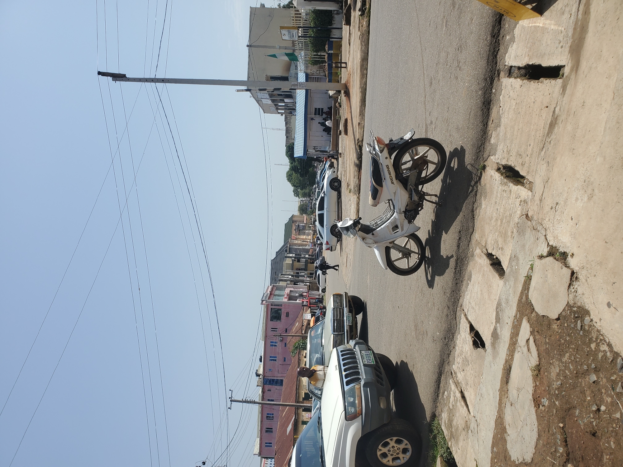 The image depicts a street scene in Jos, Nigeria. In the foreground, a white Jincheng motorcycle is parked on the right side of the road, casting a shadow. To the left, a white Jeep Grand Cherokee is also parked. The road surface is asphalt, with a concrete drainage system in the immediate foreground that appears damaged in places.
Further down the street, a man in a yellow and white striped shirt is standing next to a beige Mercedes-Benz sedan. Behind these vehicles, a row of buildings lines the street, characterized by a mix of commercial and residential architecture. The buildings vary in color and style, with some featuring corrugated metal roofs and others more modern facades. A prominent pink building with multiple windows stands out among them.
In the background, more buildings and vehicles are visible, suggesting a moderately busy urban environment. A white utility pole with power lines stretches across the upper portion of the frame. The sky is clear and blue. The overall scene conveys a typical day in an urban area, with elements of commerce, transportation, and everyday life.