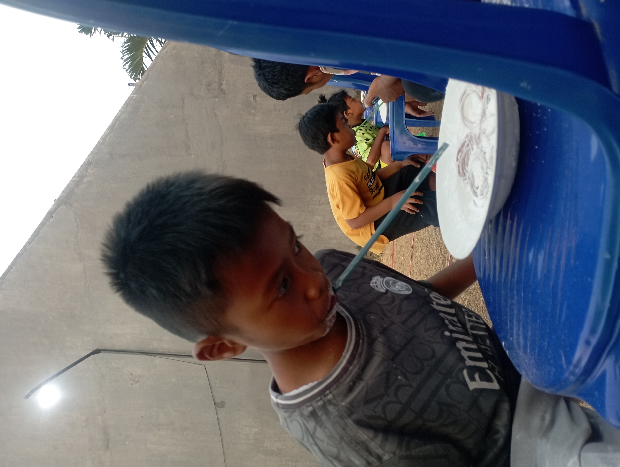 In Candirejo, Indonesia, a group of children are participating in a game. In the foreground, a young boy wearing a dark gray shirt with a white "Emirates" logo is intently focused on a white bowl placed on a blue plastic table. He is using a blue straw to attempt to pick up rings from the bowl, which is filled with white powder, likely flour or cornstarch. Several rings, possibly made of dough, are visible within the powder. The boy's face is partially obscured as he leans over the bowl, with some of the white powder clinging to his face and shirt.
Behind him, other children are also engaged in the game. One boy in a yellow t-shirt and black shorts is leaning forward, also with a straw in his mouth. Another child in a green and yellow patterned shirt is also participating, as is another individual whose face is not visible. The children are seated on blue plastic chairs. The background consists of a light-colored concrete wall and some greenery, suggesting an outdoor setting. A light fixture is mounted on the wall above and to the left of the main subject. The ground appears to be dirt or gravel. The scene is captured in daylight, with a neutral, overcast sky.