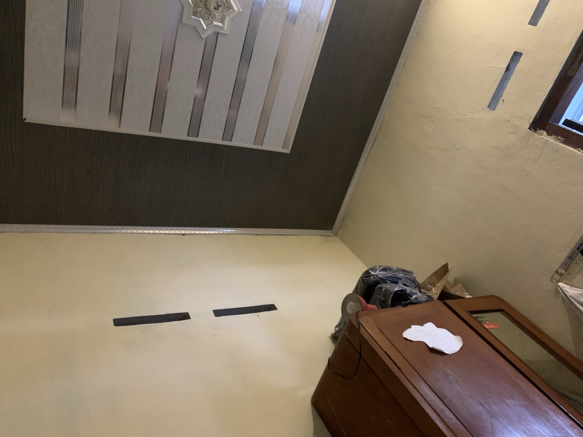 The image captures an interior scene, likely a room, oriented such that the floor appears at the top and the ceiling at the bottom. The walls are painted a light cream color, with a dark brown textured wallpaper section on the right. A white patterned ceiling, featuring horizontal stripes in shades of gray and beige and a decorative starburst fixture, is visible.
In the foreground, a dark brown wooden cabinet with a glass-fronted drawer is positioned on the left. A white, irregular-shaped object, possibly a sticker or a cut-out, is affixed to the cabinet door. To the right of the cabinet, a roll of red and gray tape is partially visible, along with some items wrapped in clear plastic.
On the cream-colored wall, two dark rectangular strips are vertically oriented. Towards the bottom of the image, a portion of another cabinet or furniture piece with a dark wooden frame is visible, with a glimpse of a window or opening. The overall lighting suggests an indoor environment, possibly with artificial light sources. The image does not provide any specific geographical indicators to pinpoint the location within Rejoso, Indonesia.