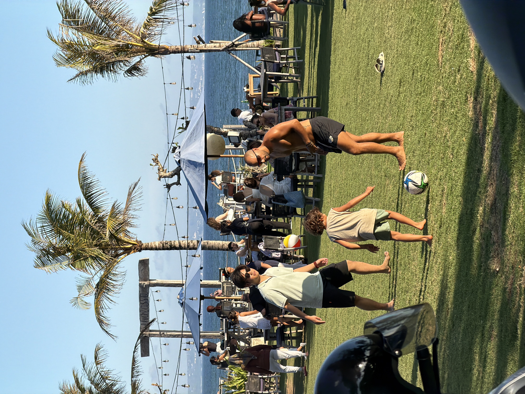 The image depicts a beachfront scene in Mengwi, Indonesia, likely at a resort or restaurant with outdoor seating. The setting features a grassy area in the foreground, leading to a beach with the ocean visible in the background. Several palm trees are scattered throughout the scene. In the foreground, two individuals, a man and a child, are playing with a soccer ball. The man is bare-chested, wearing black shorts, and the child is wearing a light-colored t-shirt and shorts. Behind them, numerous people are seated at tables under blue umbrellas, and some are standing. A wooden sign with the word "CHALASSA" is visible, along with strings of lights. The overall atmosphere suggests a relaxed, daytime gathering by the sea.