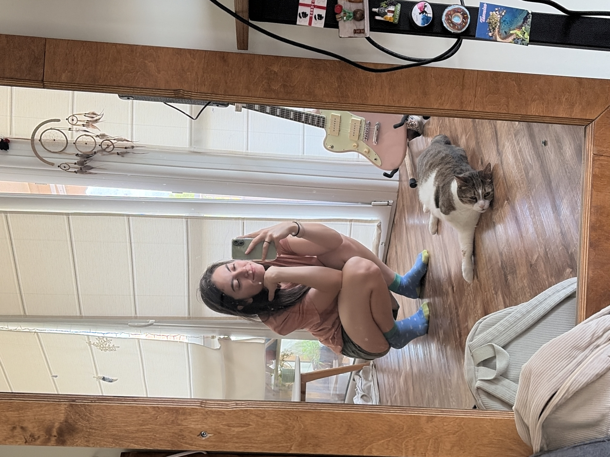 A young woman is captured in a mirror selfie, squatting on a wooden floor. She is wearing a pink t-shirt, dark shorts, and blue socks with yellow accents. She holds a smartphone in her right hand, angled to capture her reflection. Her left hand rests on her chin, and she smiles gently at the camera.
Beside her, a tabby and white cat lies stretched out on the floor, looking towards the camera with its head tilted. The background reveals a room with large glass doors leading outside, covered by white blinds. A pink electric guitar stands in a guitar stand near the doors. Decorative items, including a dreamcatcher and various magnets, are visible on a shelf to the right. A light-colored backpack is placed on the floor in the foreground. The overall environment appears to be a casual indoor setting.