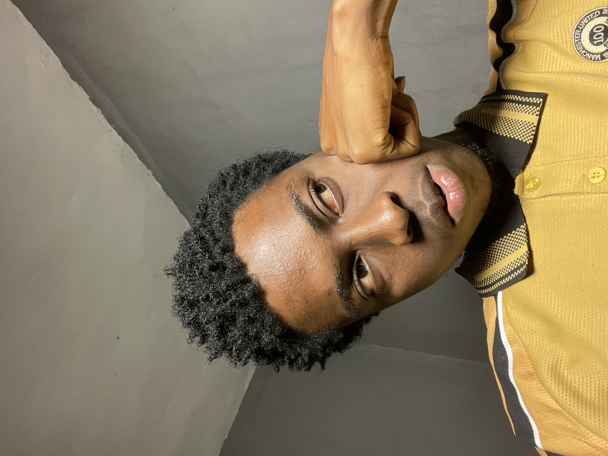 The image shows a close-up, low-angle, waist-up shot of a young Black male. He is indoors, in what appears to be a dimly lit room with a grey, textured wall that meets the ceiling at an angle. The man has dark skin, short, curly black hair, and his right fist is resting against his right cheek. He is wearing a yellow polo shirt with black and white stripes across the shoulders and a black collar with a yellow striped pattern. A circular Manchester United crest with the number 100 is visible on the right side of his chest. His eyes are looking to his left, and his lips are slightly parted. The lighting suggests it could be nighttime, with the light source likely coming from above and to his left. There are no visible clues in the image to suggest a specific location in Nkpor, Nigeria.