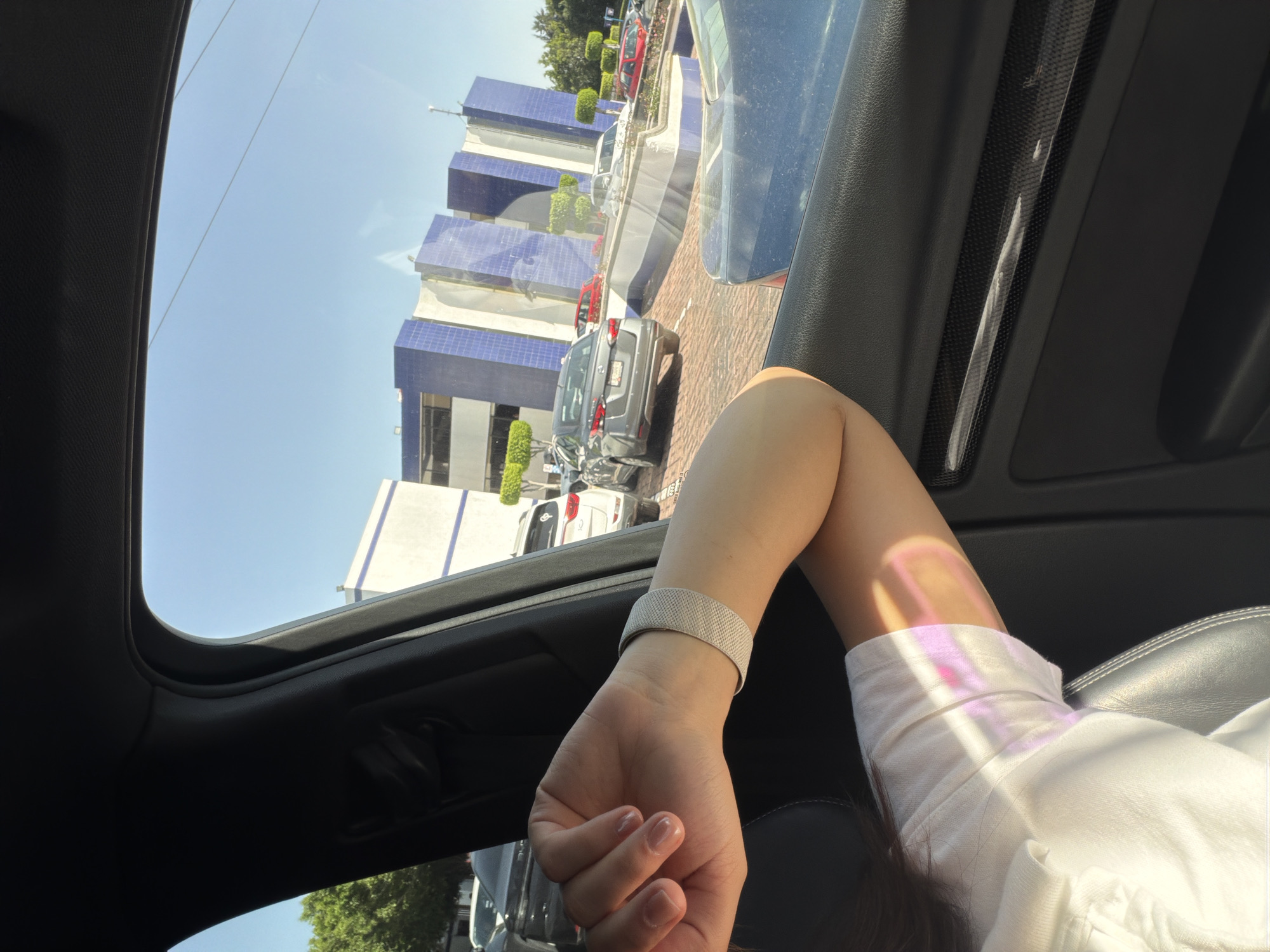 The image presents a view from inside a car, looking out through an open sunroof and a side window. In the foreground, the right arm and shoulder of a person are visible in the lower right, wearing a white short-sleeved top. A light-colored mesh watch band is on their wrist. A notable detail is the colorful light refraction, showing pink, purple, and yellow hues, cast onto the person's arm and shirt, likely from sunlight passing through the car's glass.
Outside the car, a clear, bright blue sky is prominent in the upper left. The exterior scene depicts an urban or suburban environment in San Bernardino Tlaxcalancingo, Mexico. Several modern buildings with striking blue and white facades, some with reflective surfaces, are visible. These structures appear to be commercial or office buildings. A paved area, possibly a parking lot with a brick or cobblestone pattern, hosts various parked or slowly moving vehicles, including a grey SUV, a blue car, and parts of a red car. Green trees and well-maintained bushes add to the landscaping.
The bright sunlight and absence of clouds indicate a clear weather day, likely mid-morning or afternoon. The scene conveys a calm, stationary moment or slow travel from within the car. No discernible text is clearly visible on any of the buildings or vehicles.