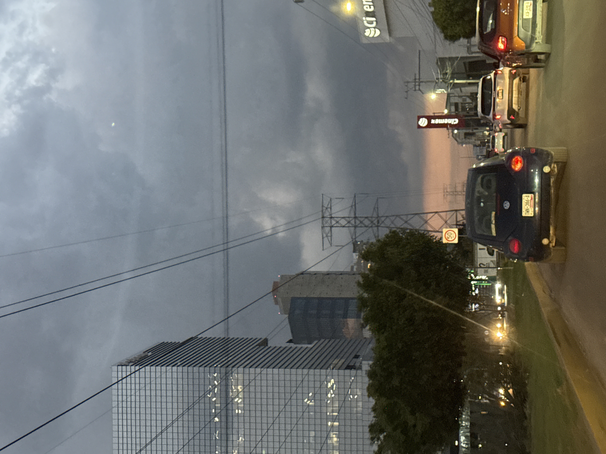 The image shows a street scene in Puebla, Mexico at dusk. A tall, modern office building with a gridded glass facade is visible in the lower left. Behind it and slightly to the right is another taller building with a rectangular base and a smaller, cantilevered section above. Overhead power lines run across the sky.
Several cars are stopped in traffic on the right side of the frame. A dark blue Volkswagen Beetle is closest, with its license plate (TWN-386-B) visible. Behind it are a white SUV and an orange Jeep. Signs for Cinépolis and Citibanamex are visible. A speed limit sign indicating 50 km/h is present near the Beetle. Trees line the street on the right, and a large electrical transmission tower is located among the trees. The sky is overcast with hints of pink and orange near the horizon.