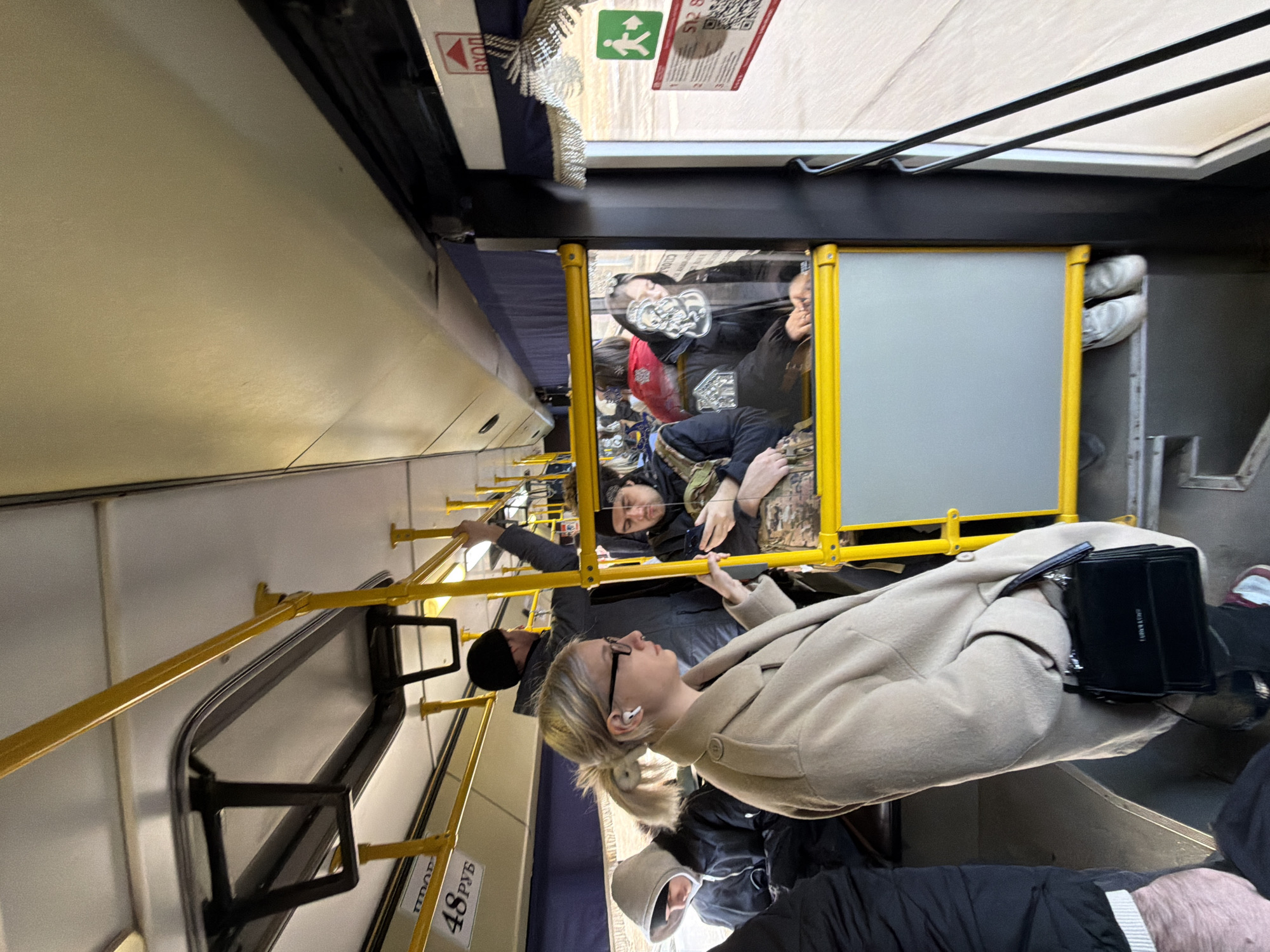 The image shows the interior of a bus, likely in Krasnoyarsk, Russia, based on the Cyrillic text visible. Passengers are seated and standing inside the moving bus. Yellow handrails are visible. People are wearing winter clothing. One woman in the foreground wears a tan coat and glasses and has a black bag. A fare sticker is visible.