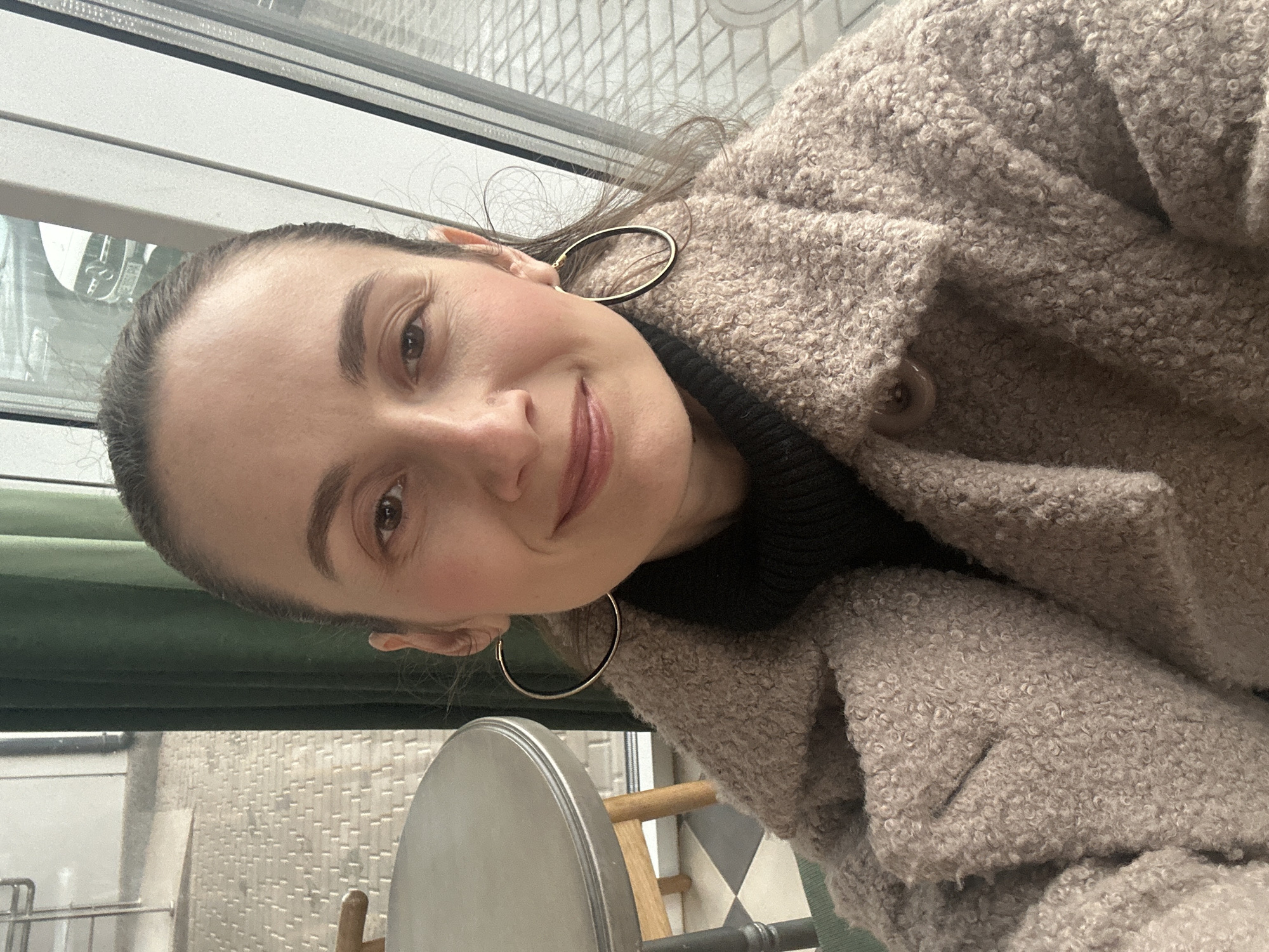 A woman with dark hair pulled back captures a close-up, chest-up selfie indoors, likely in a cafe or restaurant in Kryvyi Rih, Ukraine. She displays a soft, pleasant smile, looking directly at the camera with brown eyes. She wears large, thin gold hoop earrings and is dressed in a light brown or beige textured coat, possibly made of boucle or teddy fabric, layered over a black high-necked top. A button is visible on the lapel of her coat.
The background, viewed through a large window, shows an urban environment with light-colored, patterned building facades and hints of a tiled street or pathway. Inside, below the window, a dark green blind or panel is present. Further into the indoor space, a large, reflective silver circular object is seen in the lower left, possibly part of a table or serving tray. Adjacent to it are elements of wooden furniture and a distinctive floor with a black and white or dark and light grey checkered pattern. The natural light suggests it is daytime. The image conveys a relaxed and content mood. No discernible text is visible.