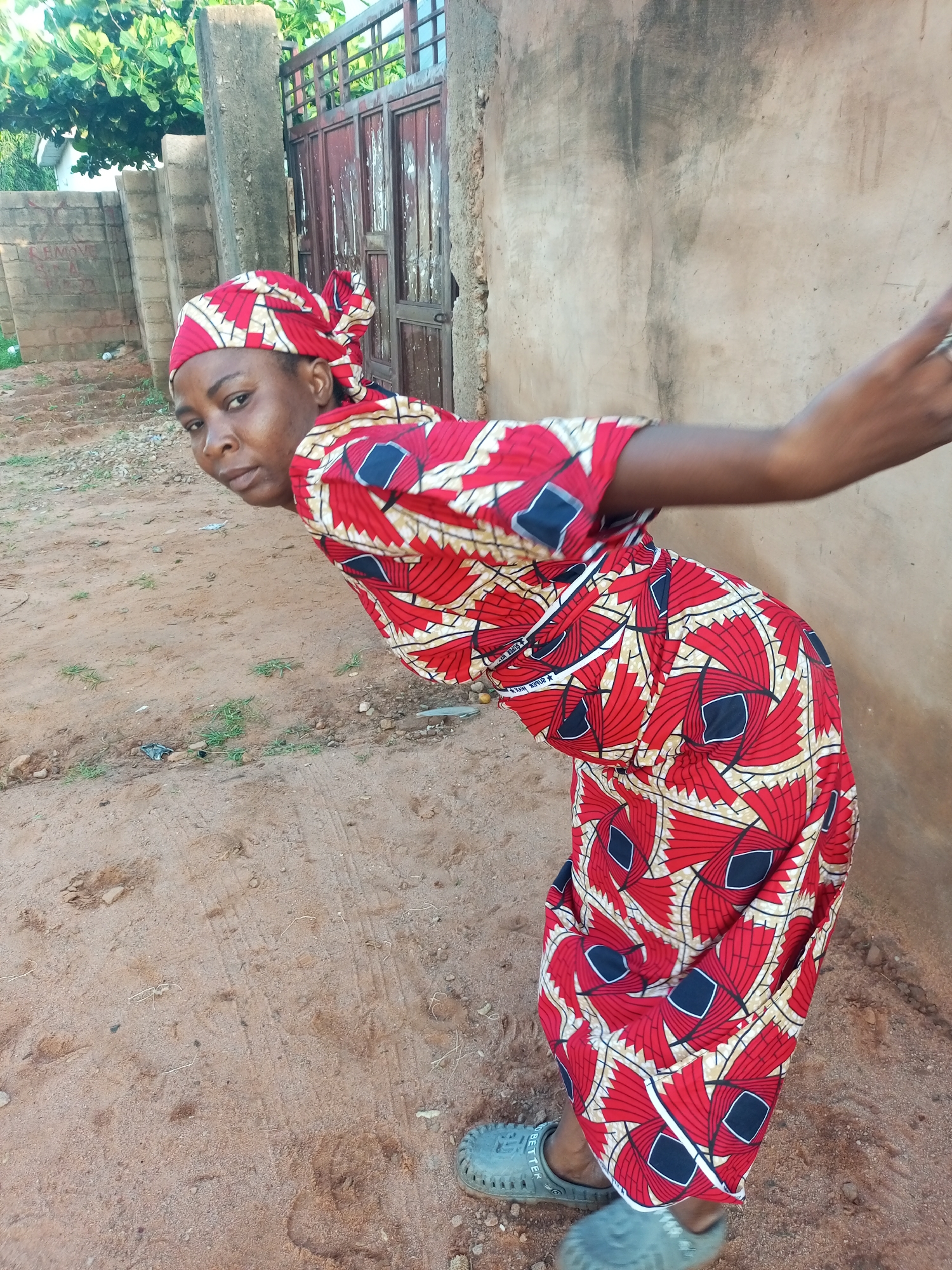 A woman wearing a red and white patterned outfit and a matching headscarf is bent over, looking at the camera. She is standing on a dirt ground in front of a beige wall. In the background, there is a metal gate and some trees. The scene appears to be outdoors in Tempure, Nigeria.