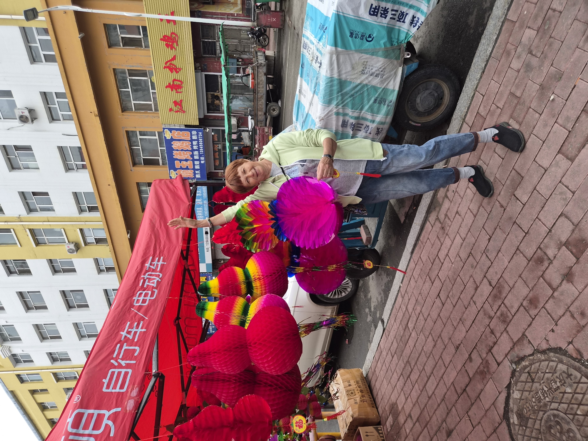 A woman with short red hair smiles as she stands on a brick sidewalk in front of a red canopy displaying various Chinese lanterns and decorations. She is wearing a light green hooded jacket, a gray t-shirt, and blue jeans rolled up at the ankles, along with gray socks and black athletic shoes. She holds a colorful, fanned-out decoration in her left hand and has her right hand raised. Behind her, a trailer is covered with a white and blue tarp. Buildings with yellow and white facades are visible in the background, along with street signs written in Chinese. The scene appears to be a street market in Mishan, China.