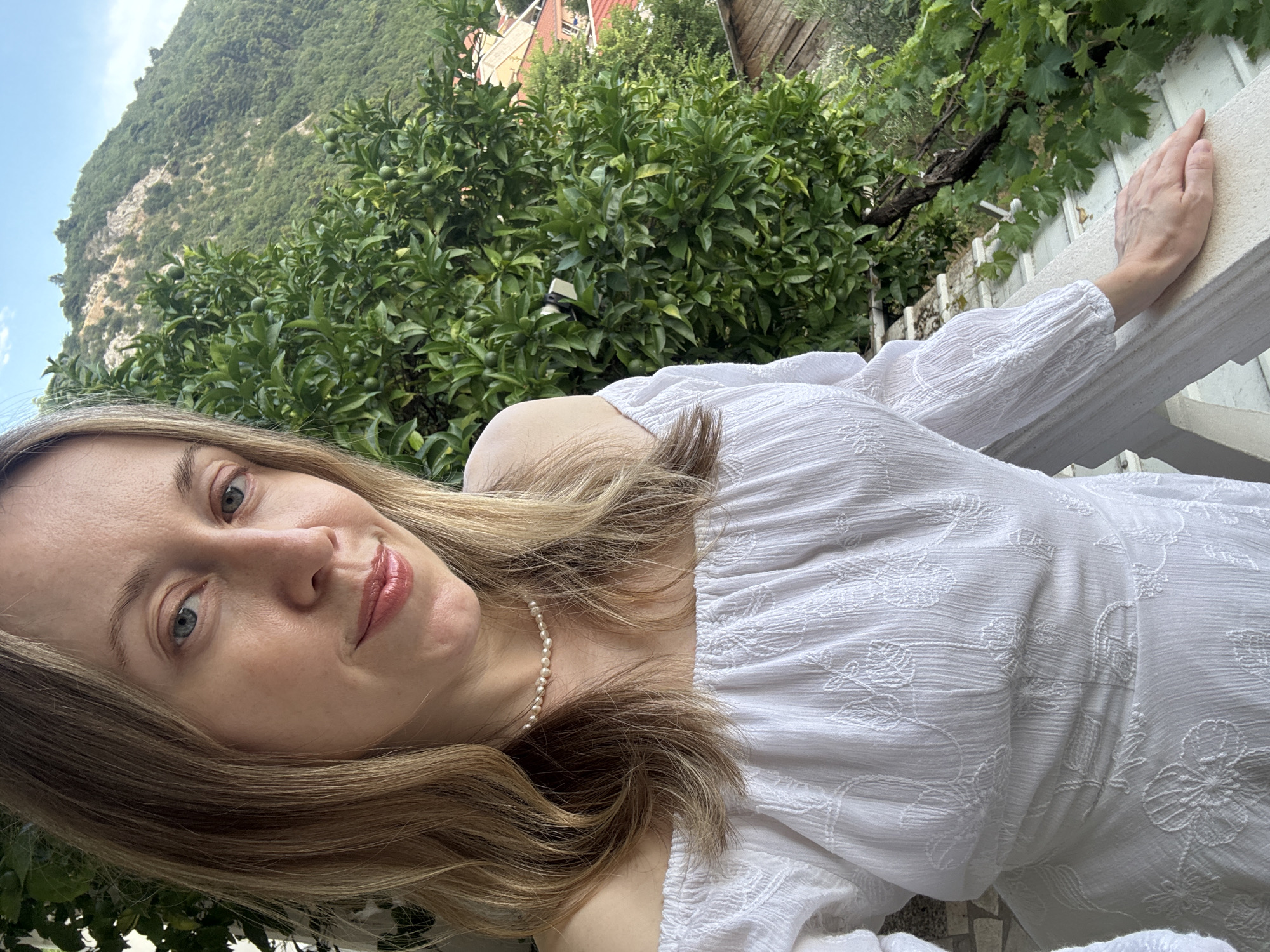 The media file is a self-portrait of a woman with long blonde hair, wearing a white off-the-shoulder dress with an embroidered floral pattern and a pearl necklace. She has light blue eyes and is smiling gently. Her right hand rests on a white railing.
The background is a lush green landscape, with a dense cluster of trees in the middle ground, possibly citrus trees given the small green fruits visible. Behind the trees, a steep, vegetated mountain rises under a clear blue sky. A hint of buildings with red roofs can be seen at the top of the mountain. The scene suggests a warm, sunny day, likely in the late morning or early afternoon.
The woman appears relaxed and content, enjoying the view. The setting, with the mountain and greenery, suggests an outdoor location in a hilly or mountainous coastal area. The presence of a railing indicates a balcony or terrace. Considering the provided location context of Budva, Montenegro, this scene likely depicts a view from a residence or hotel overlooking the Adriatic coast and its surrounding natural beauty. There is no visible text in the image.