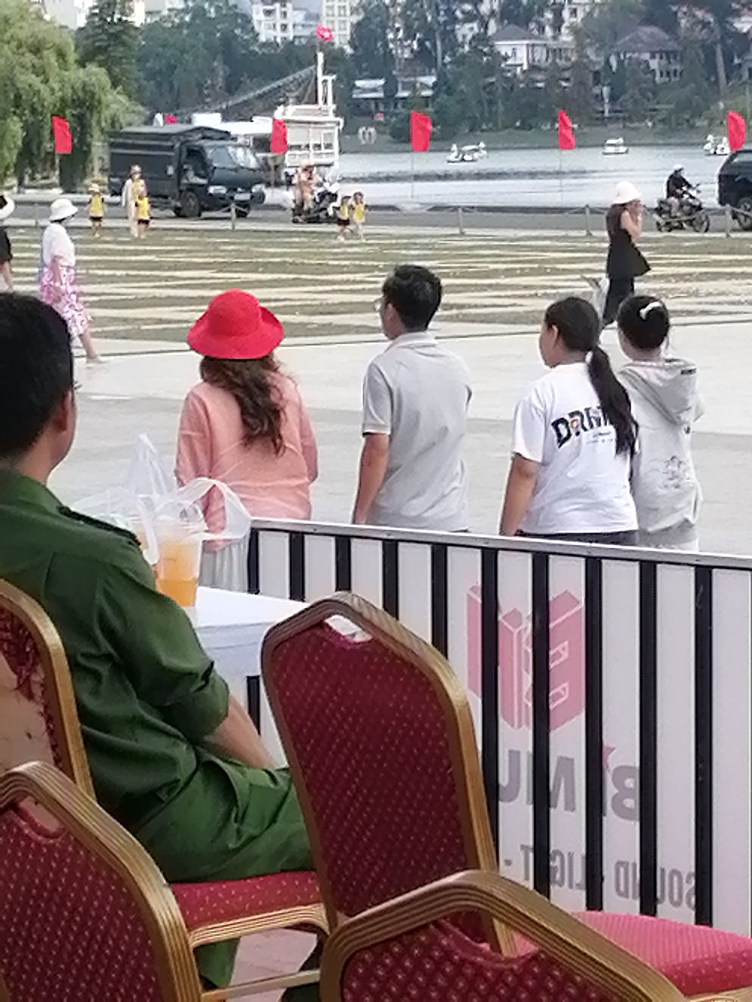 The image captures a scene near Xuan Huong Lake in Da Lat, Vietnam, likely during an event. In the foreground, two individuals are seated on red upholstered chairs with gold trim. The person on the left wears a green uniform. The person on the right, partially visible, wears a pink shirt and a red hat. Between them sits a table with two cups of orange liquid and plastic bags.
Behind them, a crowd is gathered. A woman in a pink and white patterned dress walks towards the left. A person in a red hat and pink shirt is visible. Further back, three young people are seen walking. One wears a white t-shirt with "DARA" printed on it, and another wears a light-colored hooded sweatshirt.
In the middle ground, a black truck is parked, and a motorcycle with a rider is in motion. Red flags are displayed on poles. Across the water, several boats and people can be seen on what appears to be a calm lake. The background shows buildings and lush greenery, characteristic of Da Lat's landscape. The overall atmosphere suggests a public gathering or festival.