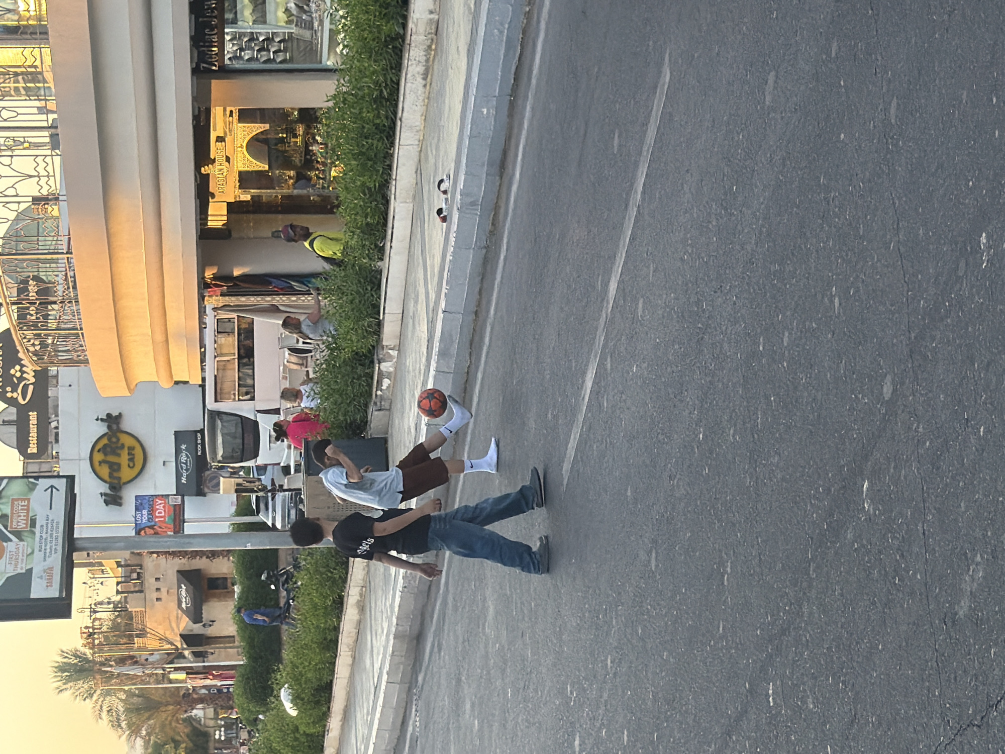 In Sharm El Sheikh, Egypt, two boys are playing football on the asphalt road bordering a sidewalk lined with lush green bushes. The boy on the left, wearing a black t-shirt and blue jeans, walks away from the viewer. The boy on the right, in a light blue shirt and brown shorts, is captured mid-action, kicking a vibrant orange soccer ball. He wears white socks and black sneakers. The background reveals a bustling street scene with buildings, including a Hard Rock Cafe, and a bus. Palm trees add to the tropical ambiance. A sidewalk features a pair of sandals near the curb. The lighting suggests it is either early morning or late afternoon, with a warm, golden glow bathing the scene.