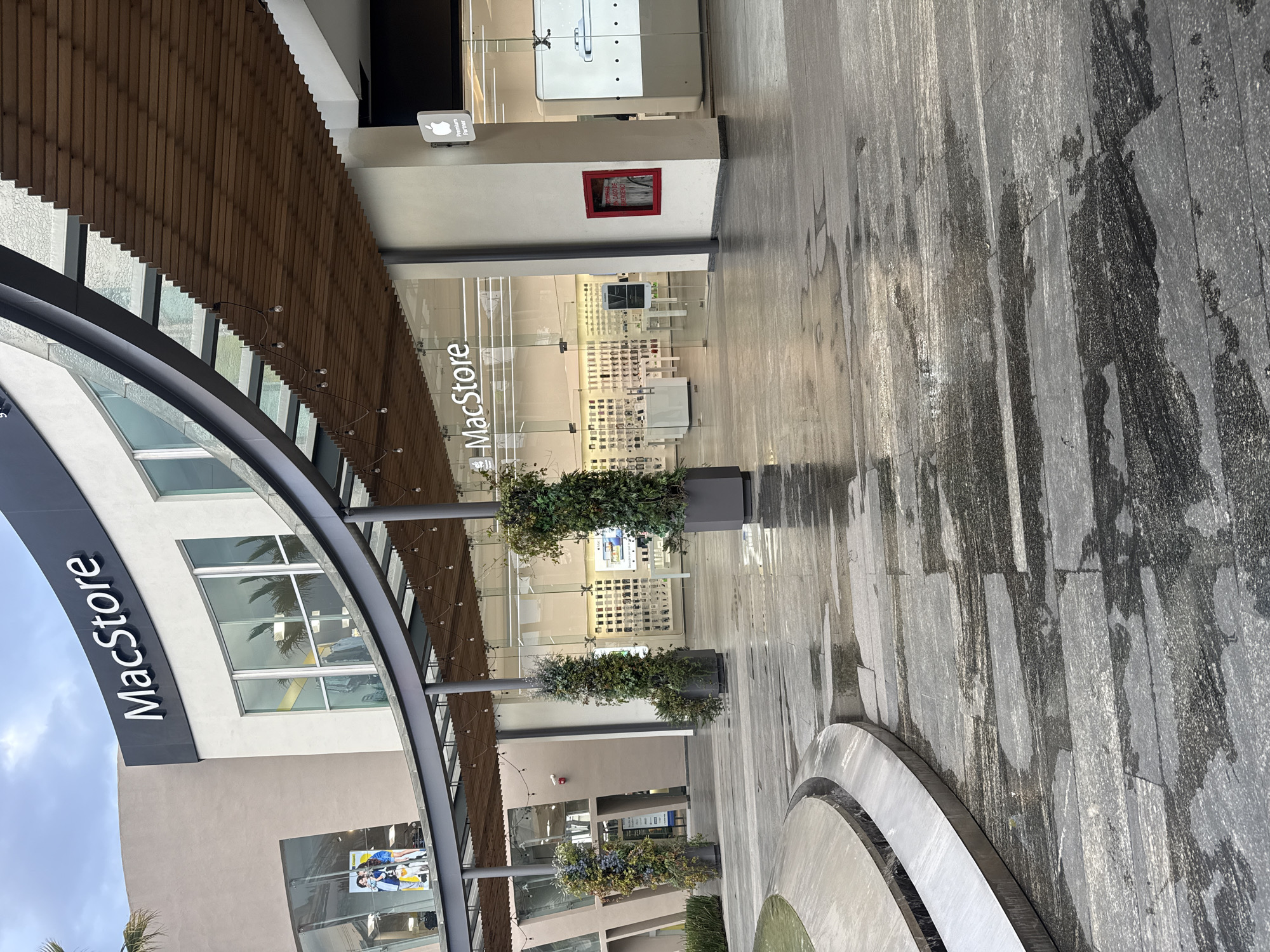 This image captures a "MacStore" retail storefront located in a contemporary, upscale shopping center. The store’s signage is prominently displayed on both the storefront and an exterior archway. Large glass windows display a variety of Apple products inside.
The scene is a pedestrian area, with patterned gray stonework. A low, circular architectural fountain element is in the foreground. Several large planters with decorative foliage are spaced along the storefront. The shot appears to be taken on an overcast day, and the wet ground suggests a recent rain. The scene is set in Puebla, Mexico.
