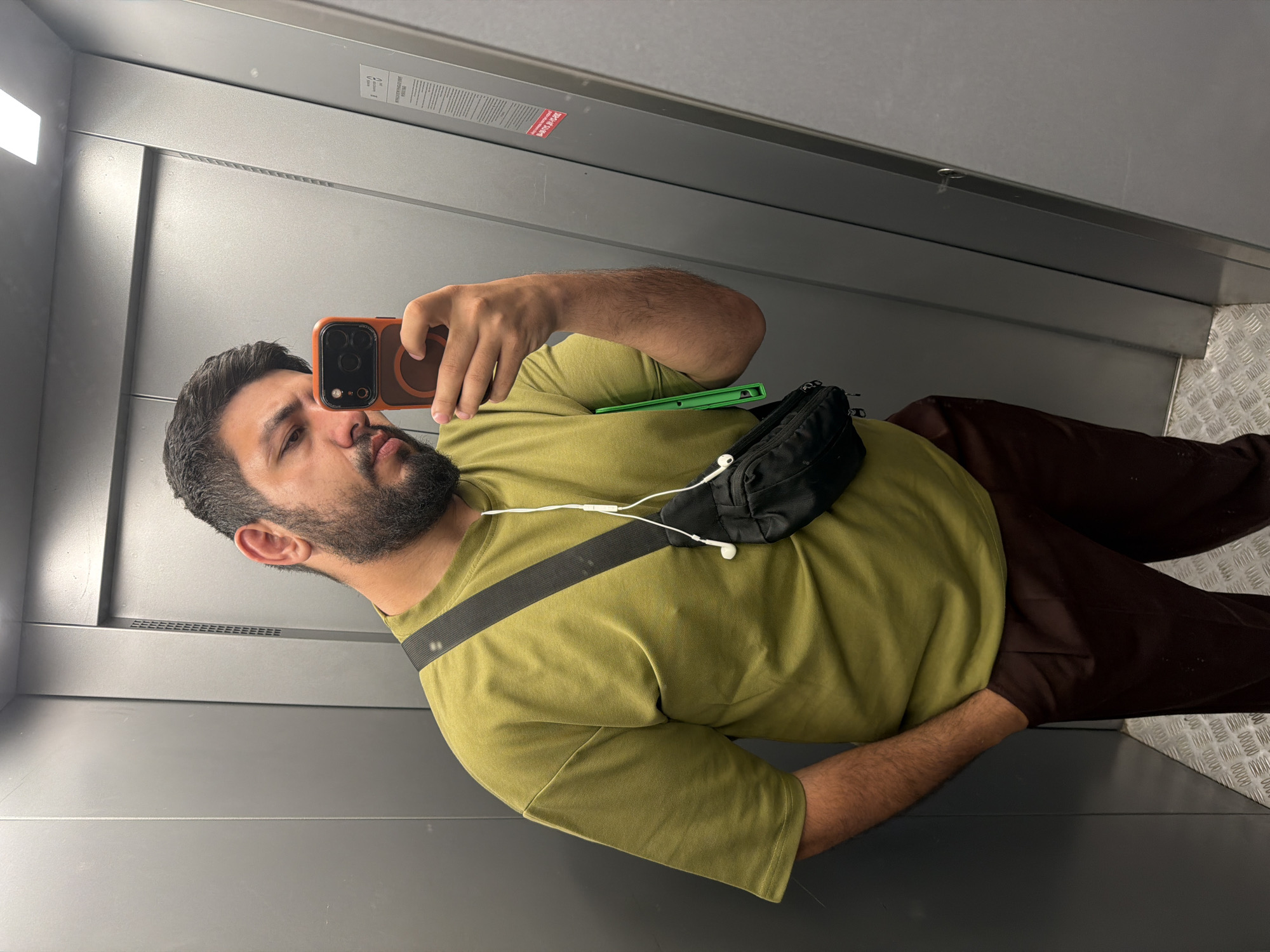 A person is taking a selfie in an elevator. The person has dark hair and a beard, is wearing an olive-green shirt, brown pants, and a black fanny pack across their chest. They are holding a cell phone in their right hand. The elevator has gray metal walls and a metal diamond-plate floor. A white light is visible at the top left of the frame. The location is possibly in Krasnoyarsk, Russia, but this is not definitively confirmed in the image.