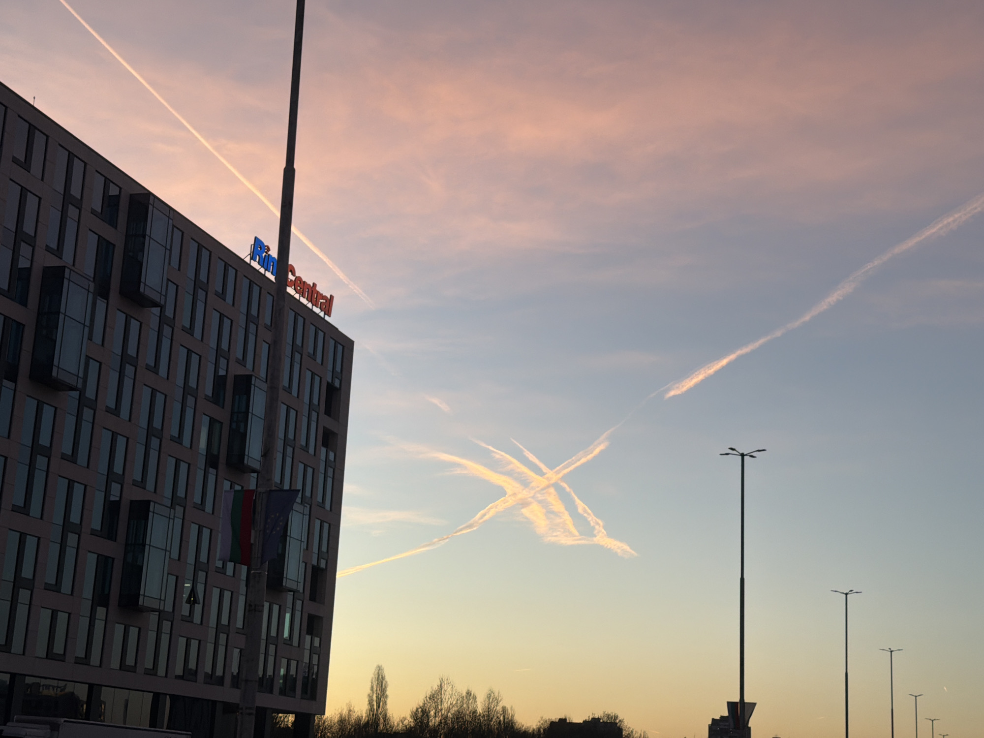 The image depicts a view in Sofia, Bulgaria, featuring a modern building with a sign reading "Ring Central" on its upper portion. The Bulgarian and European Union flags are attached to a pole near the building's entrance. The sky displays contrails from airplanes intersecting, creating an 'X' shape in the air. A row of streetlights lines the road in the foreground, with trees and other buildings visible in the distance. The sky's coloration suggests either sunrise or sunset.