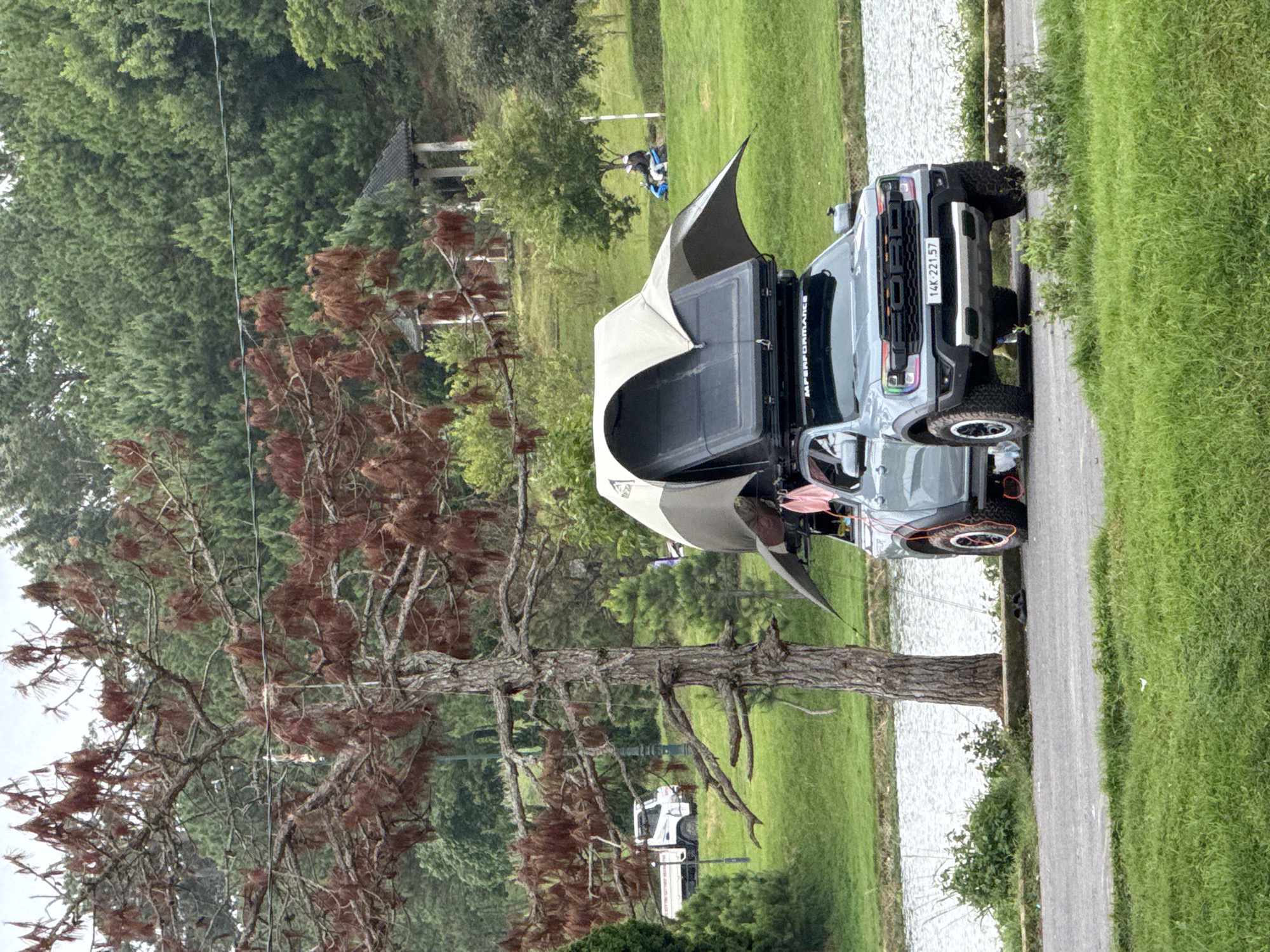 A grey Ford Ranger pickup truck with a rooftop tent is parked on a paved surface next to a body of water. The truck has a license plate that reads "14K-221.57" and "MPERFORMANCE" is visible on the roof rack. A tall tree with dry brown foliage is to the left of the truck, and a green grassy hill with a few people and a scooter is in the background. In the distance, there are more trees and some traditional Vietnamese architectural structures. The sky is overcast. The scene appears to be a park or natural area in Lâm Viên - Đà Lạt, Vietnam.
