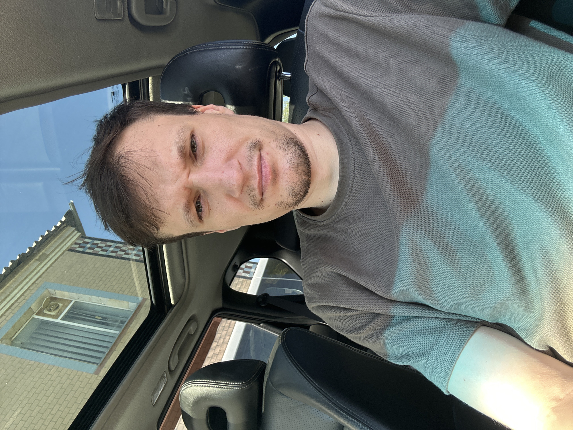 A man with brown hair and a goatee is sitting in the driver's seat of a car, looking directly at the camera with a neutral expression. The car has black leather seats and a sunroof that is open, revealing a clear blue sky. The reflection in the sunroof shows a building with light brown brickwork and a window with blue trim. The man is wearing a dark gray t-shirt. The image was taken outdoors in Shatava, Ukraine, on a sunny day, as indicated by the bright sunlight and shadows. The background shows a typical residential area with houses and vegetation.