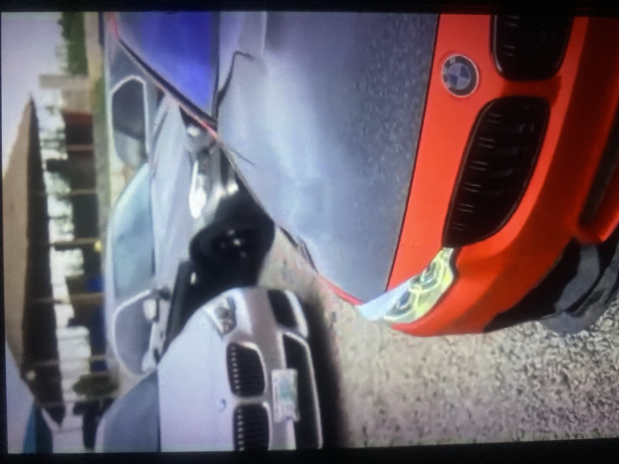 In an outdoor parking lot, a red BMW M series car with a matte black hood is parked in the foreground. To its left, a silver BMW is visible, with its front end facing the viewer. Behind these cars, other vehicles and a shaded structure with visible support beams can be seen in the background. The ground is covered with small, light-colored gravel. The image appears to be taken from a slightly elevated perspective, focusing on the front of the red BMW.