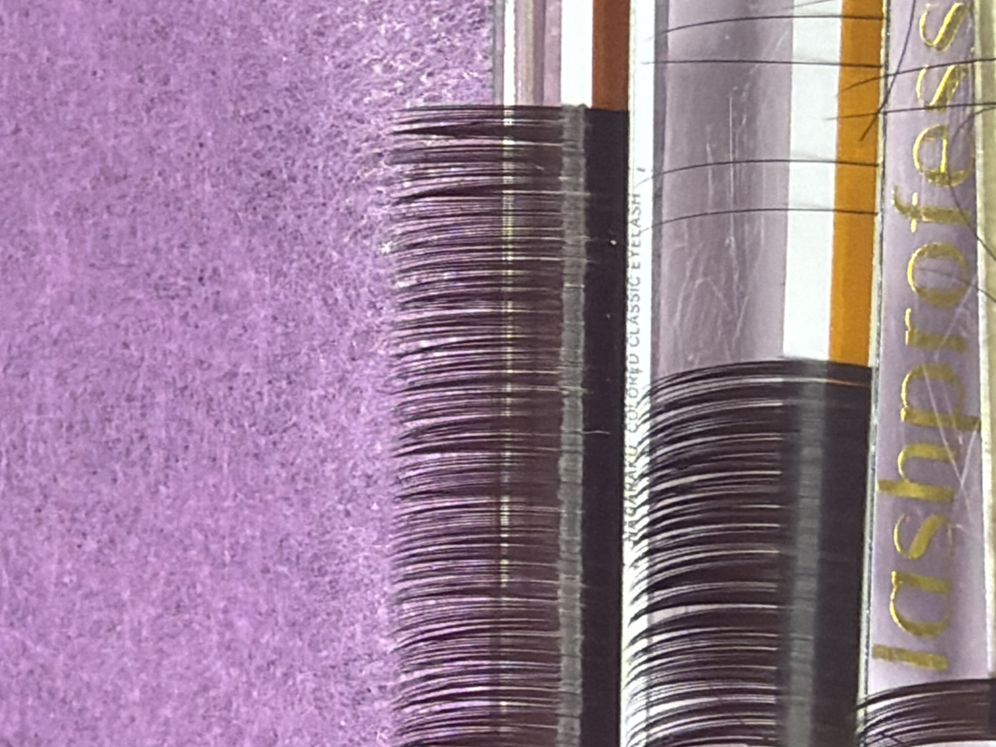 The image displays a close-up view of stacked artificial eyelashes, likely a product display or inventory. The eyelashes are arranged in neat rows, showcasing their uniformity and fine texture. A portion of the background features a textured, light purple surface, possibly fabric or a similar material.
On the right side of the image, product packaging is visible. One label has gold lettering that is partially obscured but appears to read "lash." Another label, oriented vertically, has text that is difficult to fully decipher due to the angle and resolution, but the words "COLORED EYELASH" are discernible. The packaging itself is a pale lavender color with thin, vertical lines and a bright orange stripe.
The lighting suggests an indoor setting, with no specific time of day or weather conditions evident. The focus is entirely on the product, indicating a commercial or retail context. The scene is static, with no people or discernible action taking place. The overall impression is one of a well-organized display of beauty products. The location context of Kyiv, Ukraine, does not offer any direct visual cues within the image itself.