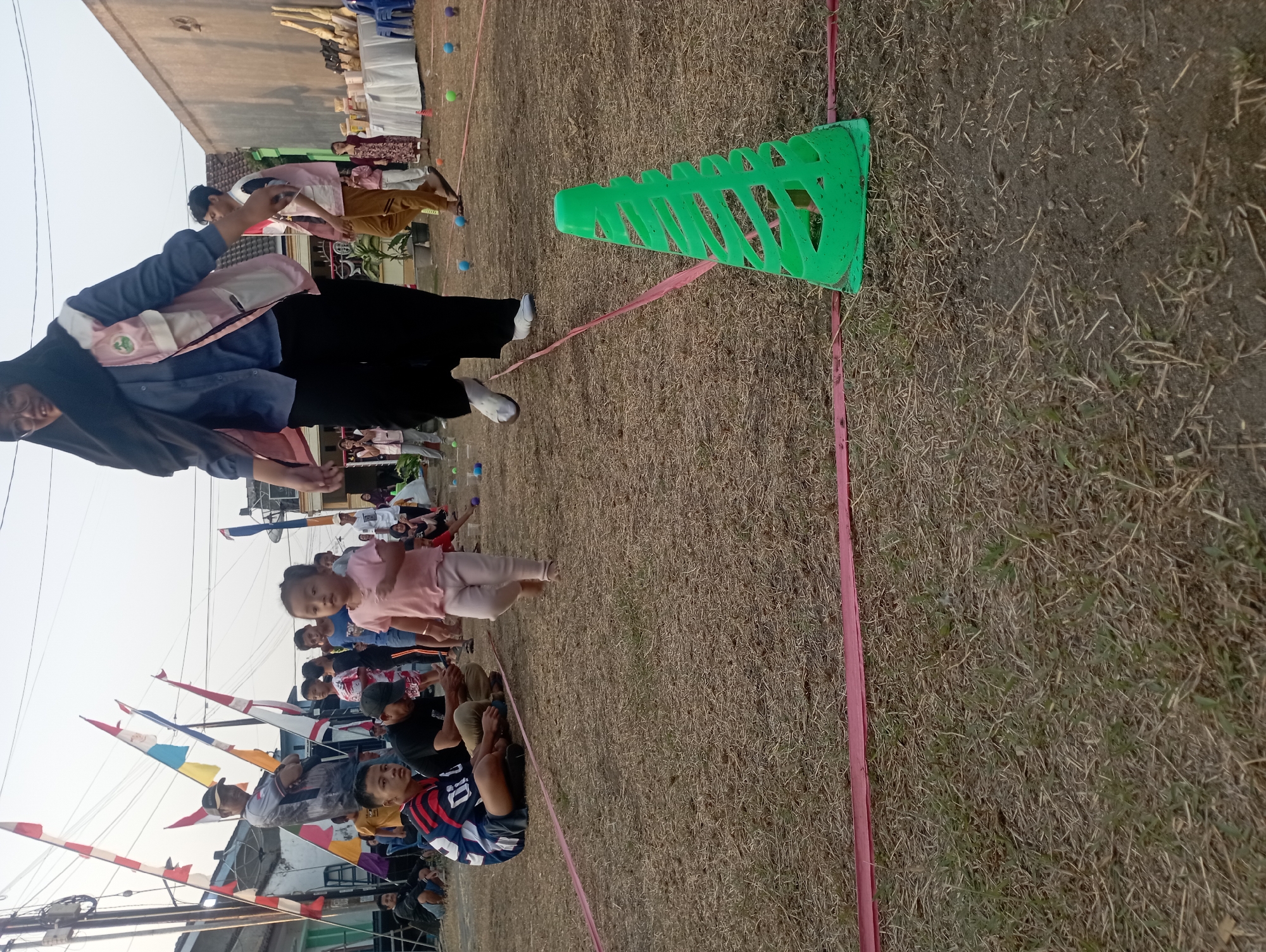 In Candirejo, Indonesia, a festive outdoor event is taking place on a dry, grassy field. A young girl, wearing a pink t-shirt and white leggings, is running towards the camera. She is part of a race, indicated by the pink ribbon marking the course and the green cone placed on the ground. In the background, a woman wearing a hijab, blue shirt, and pink vest is walking, likely a participant or organizer. Several other people, including children and adults, are gathered around, some sitting on the ground and others standing, watching the event. Flags and banners are visible, adding to the celebratory atmosphere. The scene is illuminated by the warm light of the late afternoon sun.