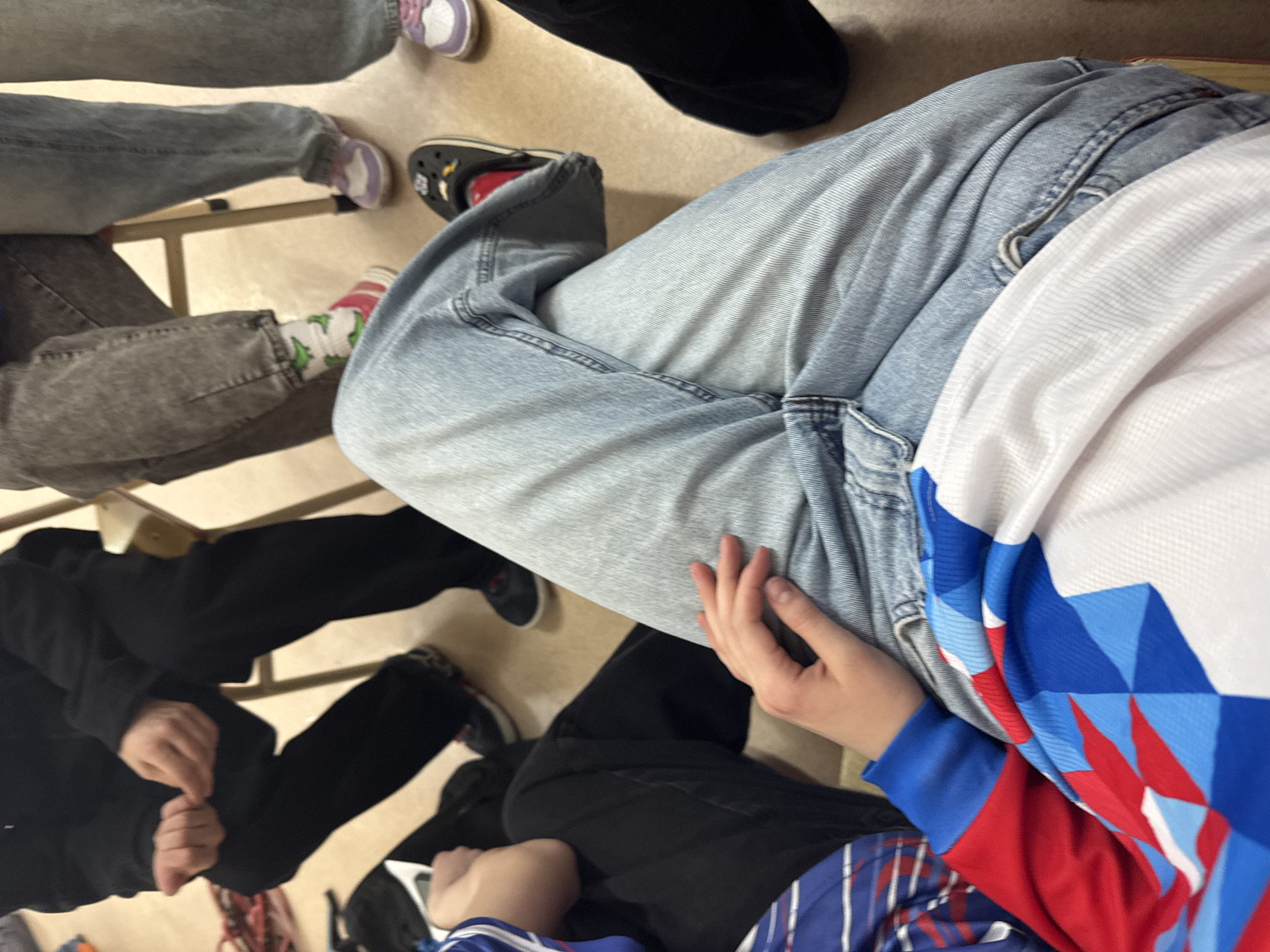 A close-up, overhead shot shows portions of multiple people sitting, possibly in an indoor setting. The floor is a light tan color. One individual wears light-wash denim jeans. Another wears black pants and a long-sleeved black jacket. A third person is wearing gray denim jeans. Another person wears black pants. One person is wearing a blue, red, and white patterned top with long blue sleeves. The feet and shoes of multiple people are visible, including a pair of purple sneakers, a pair of black Crocs with red lining, and a pair of black sneakers. One person is wearing patterned socks. This is in Usolye, Russia.