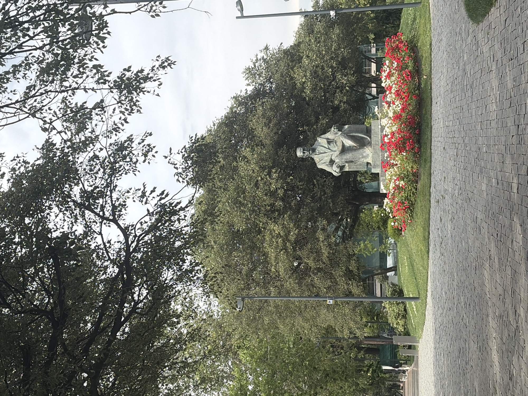 This image depicts a statue of a seated man, likely a prominent figure, positioned in a park-like setting in Tbilisi, Georgia. The statue is made of stone or bronze and is set upon a pedestal. In front of the statue, a circular flower bed bursts with red and white roses, creating a vibrant contrast against the greenery. Behind the statue, a large, mature tree with dense foliage dominates the background. The park features manicured lawns and pathways paved with cobblestones. Streetlights are visible in the background, suggesting an urban park environment. The sky is overcast, indicating a cloudy day. The overall scene is serene and suggests a public space designed for leisure and appreciation of art and nature.