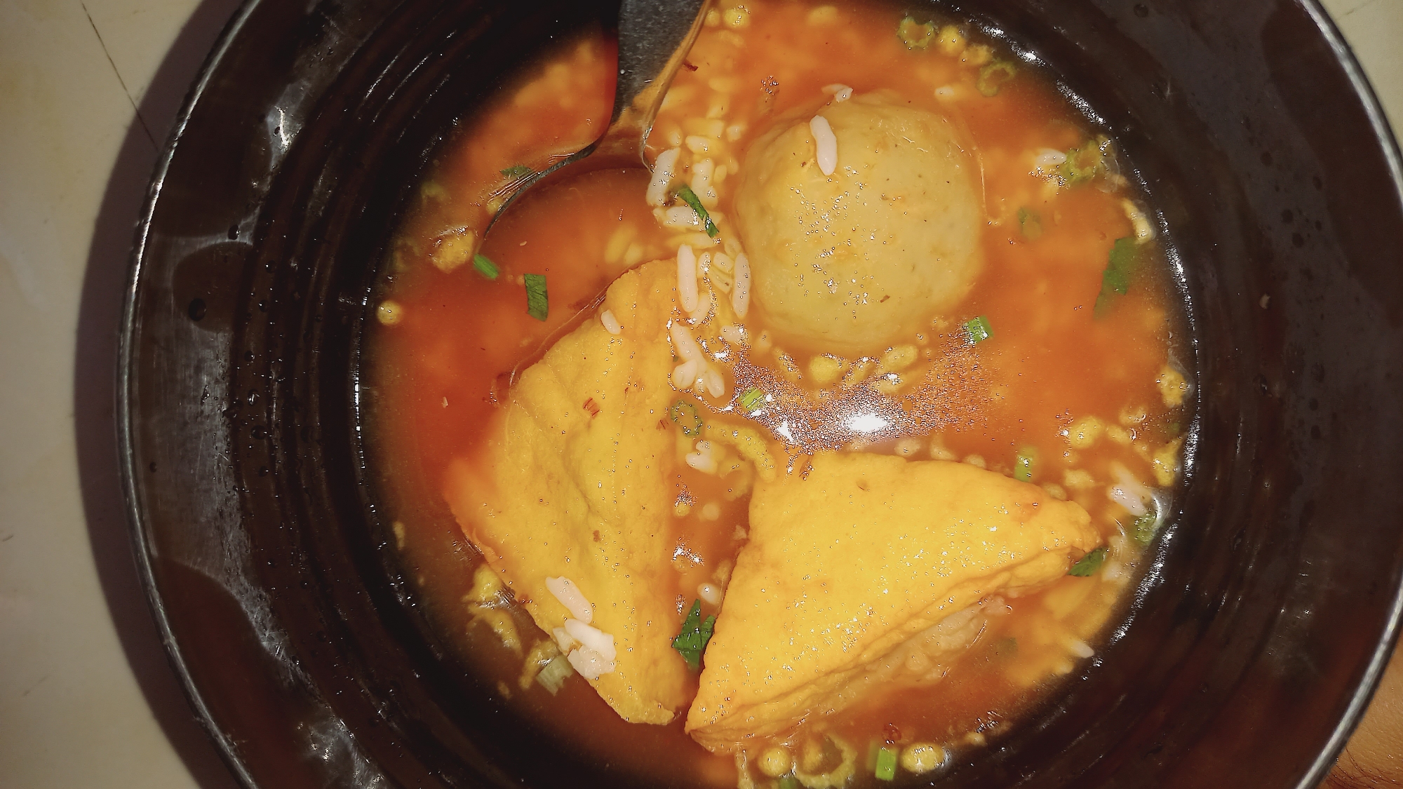 A close-up overhead shot shows a bowl of Indonesian food, likely mie tek-tek, in Leuwisadeng, Indonesia. The dish contains a rich, orange broth with visible chili oil. Floating in the broth are several pieces of fried tofu, triangular in shape, and one large, spherical meatball. Small pieces of white rice and chopped green onions are also dispersed throughout the dish. The bowl itself is dark, appearing to be made of a dark ceramic or wood material with a slightly textured inner rim. The lighting is dim, giving the scene a warm, slightly shadowy ambiance. There are no people visible in the image.