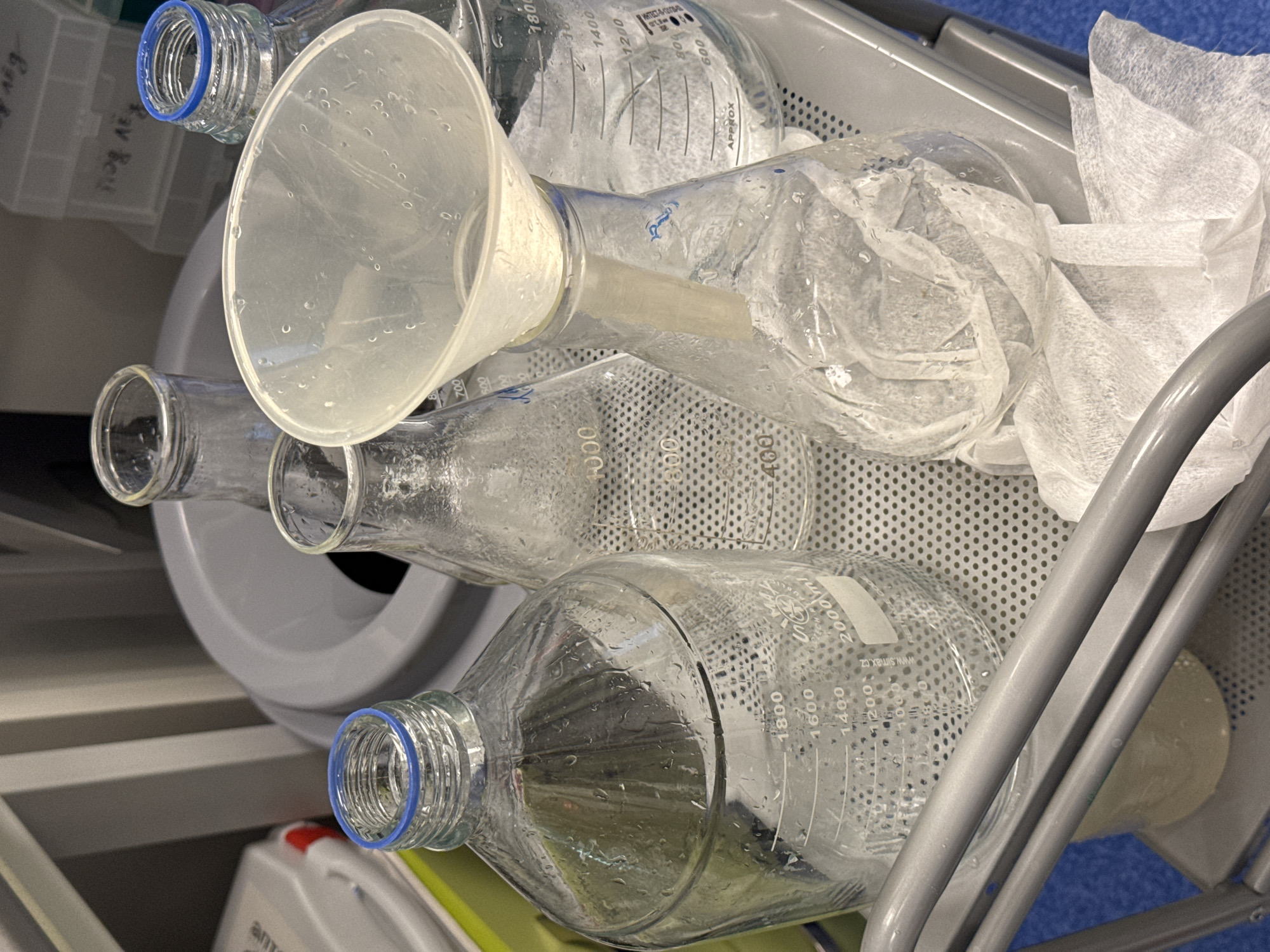 The image captures a close-up, angled view of various laboratory glassware and equipment, likely in a drying or storage cart within a scientific facility in Moscow, Russia. The primary subjects are several clear glass laboratory bottles or flasks of different capacities, a translucent plastic funnel, and crumpled white tissue paper.
All glass items, including the bottles and the funnel, are visibly wet with numerous water droplets, suggesting they have been recently washed and are in the process of drying. The bottles are lying on their sides on a perforated metal surface, which appears to be part of a cart or rack. One prominent bottle in the foreground has a blue ring around its threaded neck and volumetric markings up to "2000ml," with specific lines for "1800," "1600," "1400," "1200," and "100" milliliters. Text "www.simax.cz" is also visible on this bottle, identifying the manufacturer as a Czech glass company. Another bottle further back also shows similar markings, including "APPROX," "804," "600," and "ИНТЕСТ-А-12/".
The translucent plastic funnel, also covered in water droplets, is resting partially inside the neck of one of the bottles. To the right, crumpled white tissue or paper wipes are loosely placed on the perforated surface. The background is muted, showing hints of other lab equipment and possibly a blue floor or surface. The scene is devoid of people and there are no discernible cues for time of day or weather.