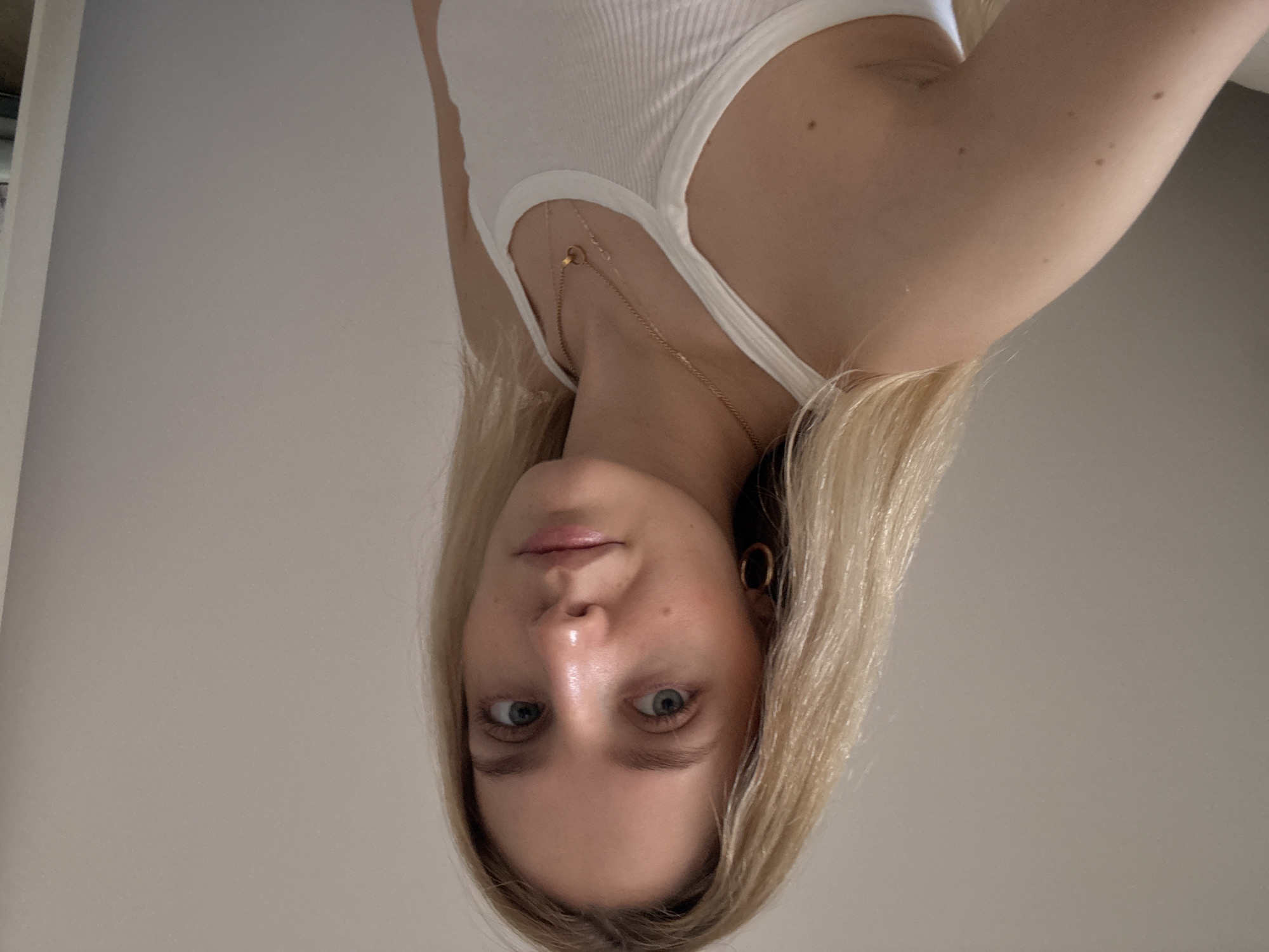 The scene depicts a female subject positioned indoors, identified as being in Mexico City, Mexico. The subject has fair skin, long, straight, blonde hair, and light-colored eyes. She is wearing a white ribbed tank top. A gold-colored hoop earring is visible on her left ear. Around her neck, she wears two thin gold-colored necklaces, one featuring a small double-ring pendant and another with a smaller circular pendant. The subject is looking directly towards the camera with a neutral expression. Her left shoulder and upper arm are prominent in the foreground. The background consists of a plain, light-colored wall, with a faint vertical line possibly indicating a corner or doorframe on the right side of the frame. No other objects or specific landmarks are discernible in the environment.