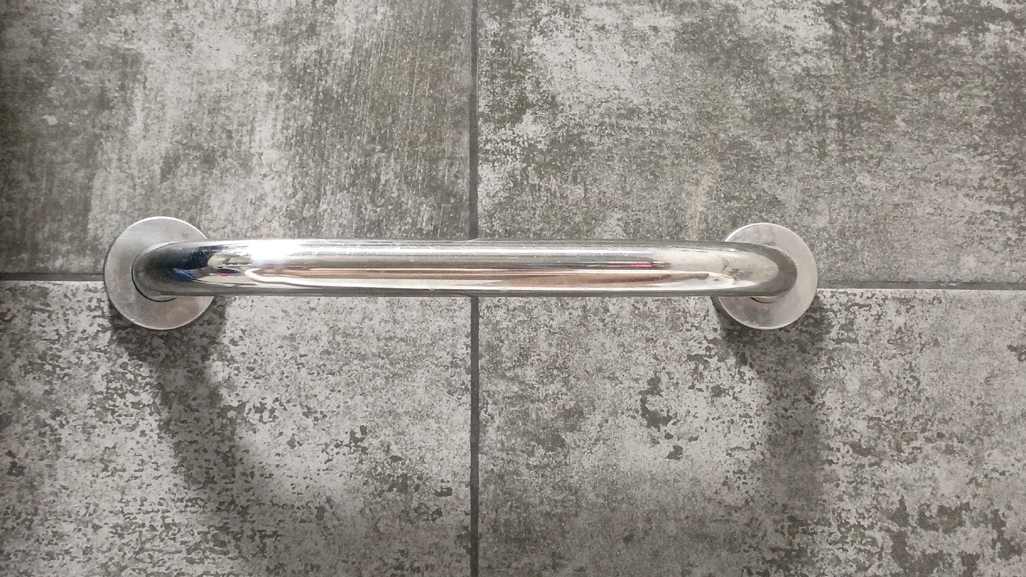 A horizontal, polished metal grab bar is mounted on a wall composed of grey, textured tiles. The grab bar is centered in the frame and is supported by circular mounting brackets at each end. The tiles exhibit a mottled pattern with variations in grey tones. Shadows are cast on the wall below the grab bar. The scene is indoors, likely in a bathroom or similar facility. The specific location in Homyel, Belarus cannot be determined from the image.