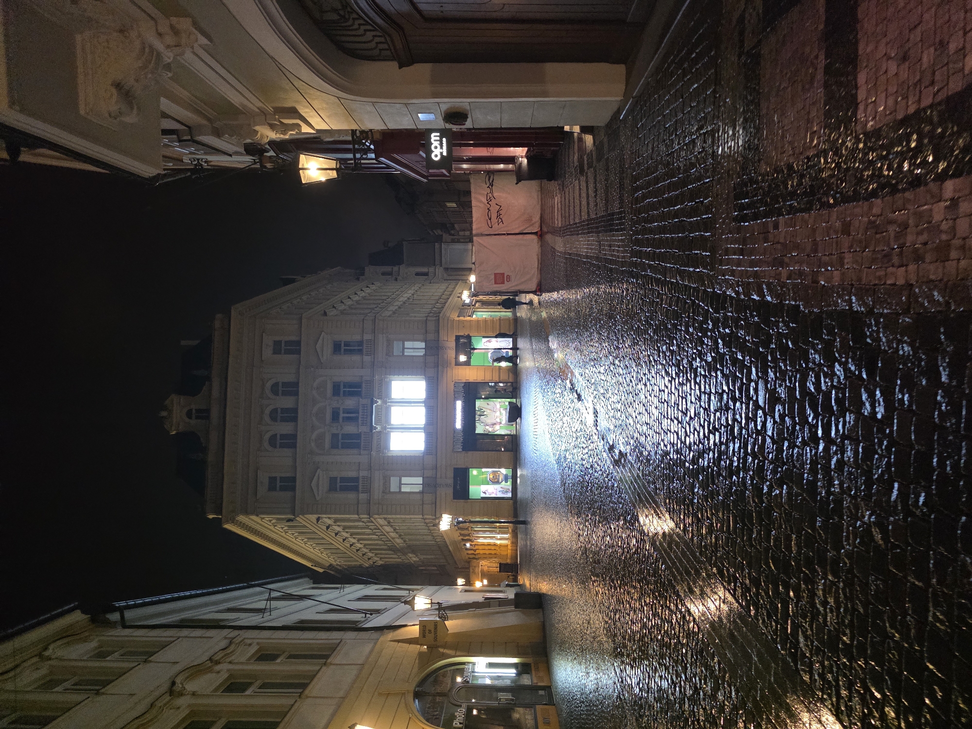 This is a nighttime, eye-level, medium-full shot of a European cobblestone street, likely in a city center, with wet surfaces reflecting the light from street lamps and building interiors. The street is paved with dark, rectangular cobblestones, some of which appear wet and reflective. On the left, there is a building with a light-colored facade, featuring arched doorways and windows. One storefront is visible, labeled "Photo" and "Souvenirs." Further down the street on the left, more storefronts are illuminated, including one with green lighting.
The center of the image is dominated by a large, multi-story building with a light-colored facade, classical architectural details, and numerous windows. The central portion of this building is brightly illuminated from within, suggesting shops or a hotel.
On the right side of the street, there is a building with a red awning and a sign reading "apm monaco." Adjacent to this is a large, white temporary construction barrier, partially covered in graffiti. A street lamp with an ornate design illuminates the area. Further down the street on the right, the building facade is a light color with architectural moldings. The street continues into the distance, with more buildings and lampposts visible. The overall atmosphere is quiet and somewhat deserted, with a few distant figures visible in the illuminated shop areas. The scene suggests it may have recently rained or is still raining, given the wet cobblestones and reflections.