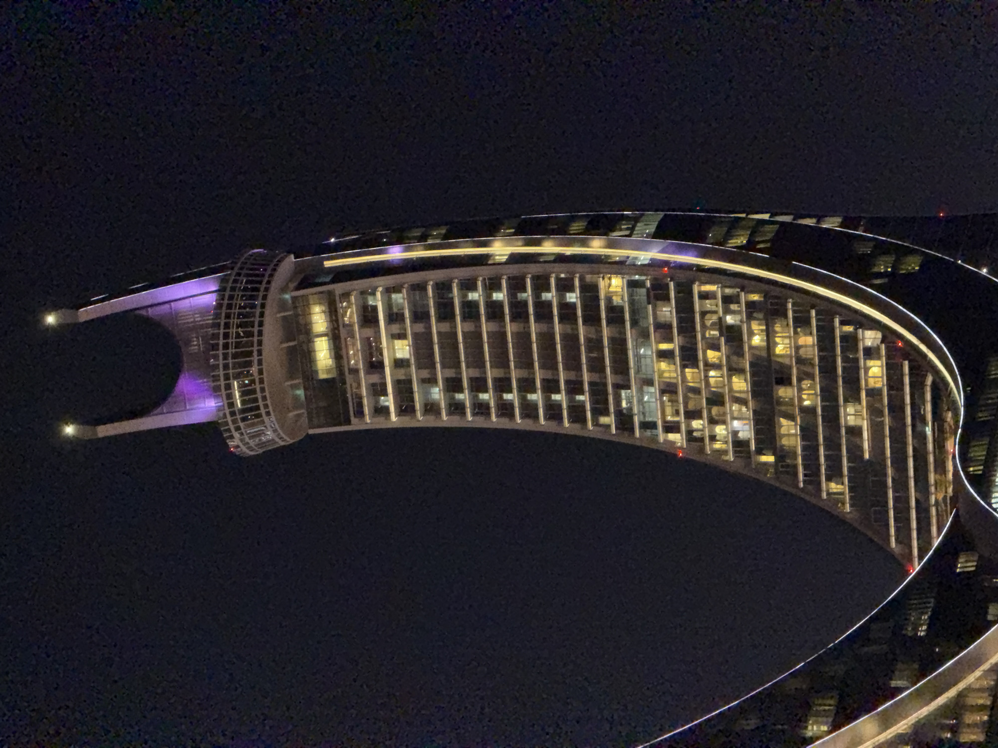 A nighttime, low-angle, eye-level shot captures a strikingly modern, curved skyscraper against a dark sky. The building's architecture is characterized by its crescent shape, with illuminated windows creating a grid pattern along its facade. A prominent circular observation deck, featuring glass railings, sits atop a section of the building. The apex of the skyscraper is crowned with a distinctive, U-shaped structure that glows with purple light, further accentuated by a bright light at its very peak. The building is outlined with bright white and yellow lights, emphasizing its unique form. The overall impression is one of a contemporary architectural marvel, likely in a major city, illuminated against the darkness of night.