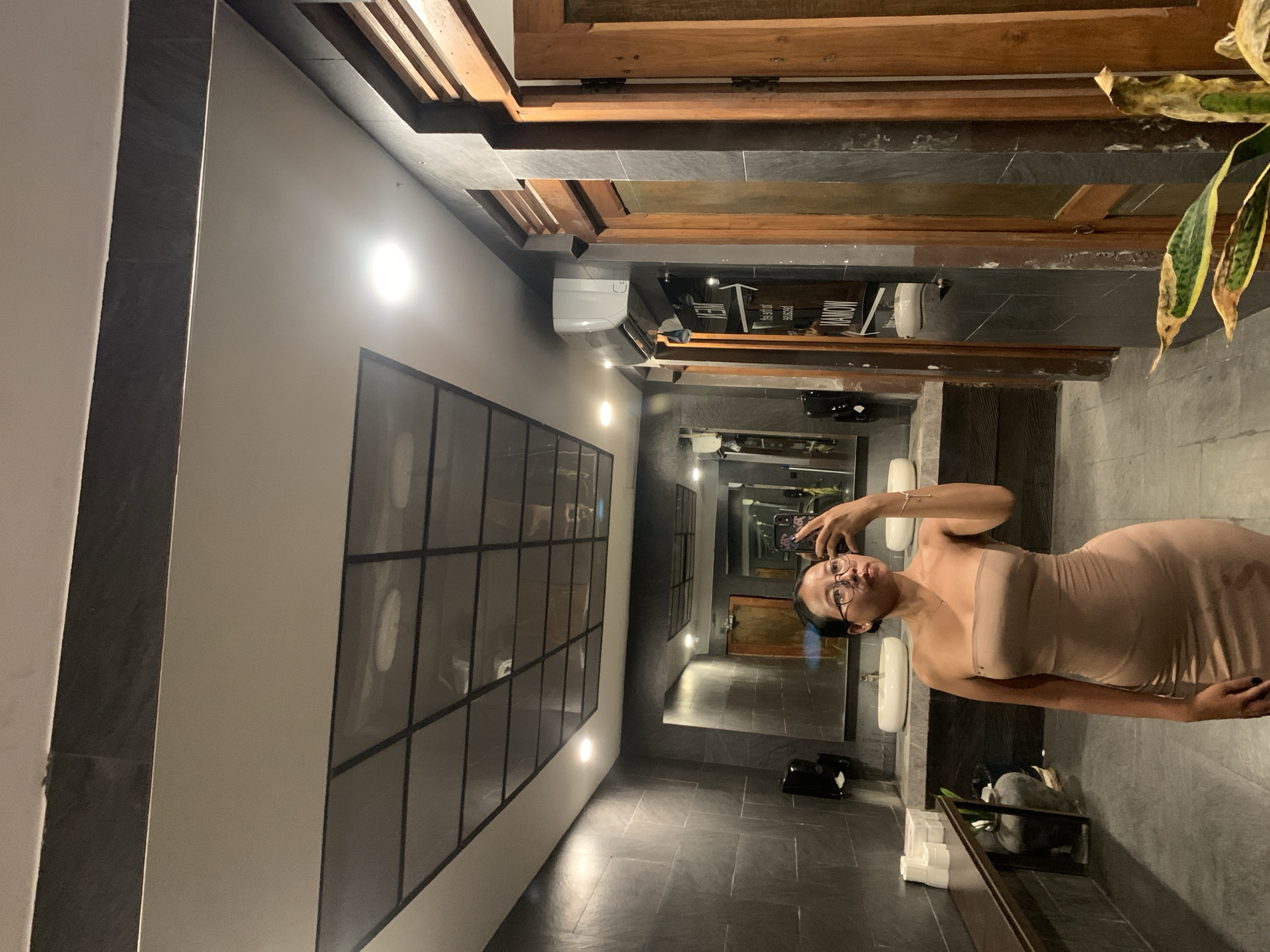 A woman with dark hair and glasses is taking a selfie in a bathroom with dark tile flooring and walls. The bathroom appears to be located in North Kuta, Indonesia. There is a large grid-patterned window on the left side of the frame. In the background, the view extends into other bathroom areas, showing mirrors and sinks. There is a wooden plank ceiling above the bathroom areas. A snake plant sits in the corner to the right of the frame.