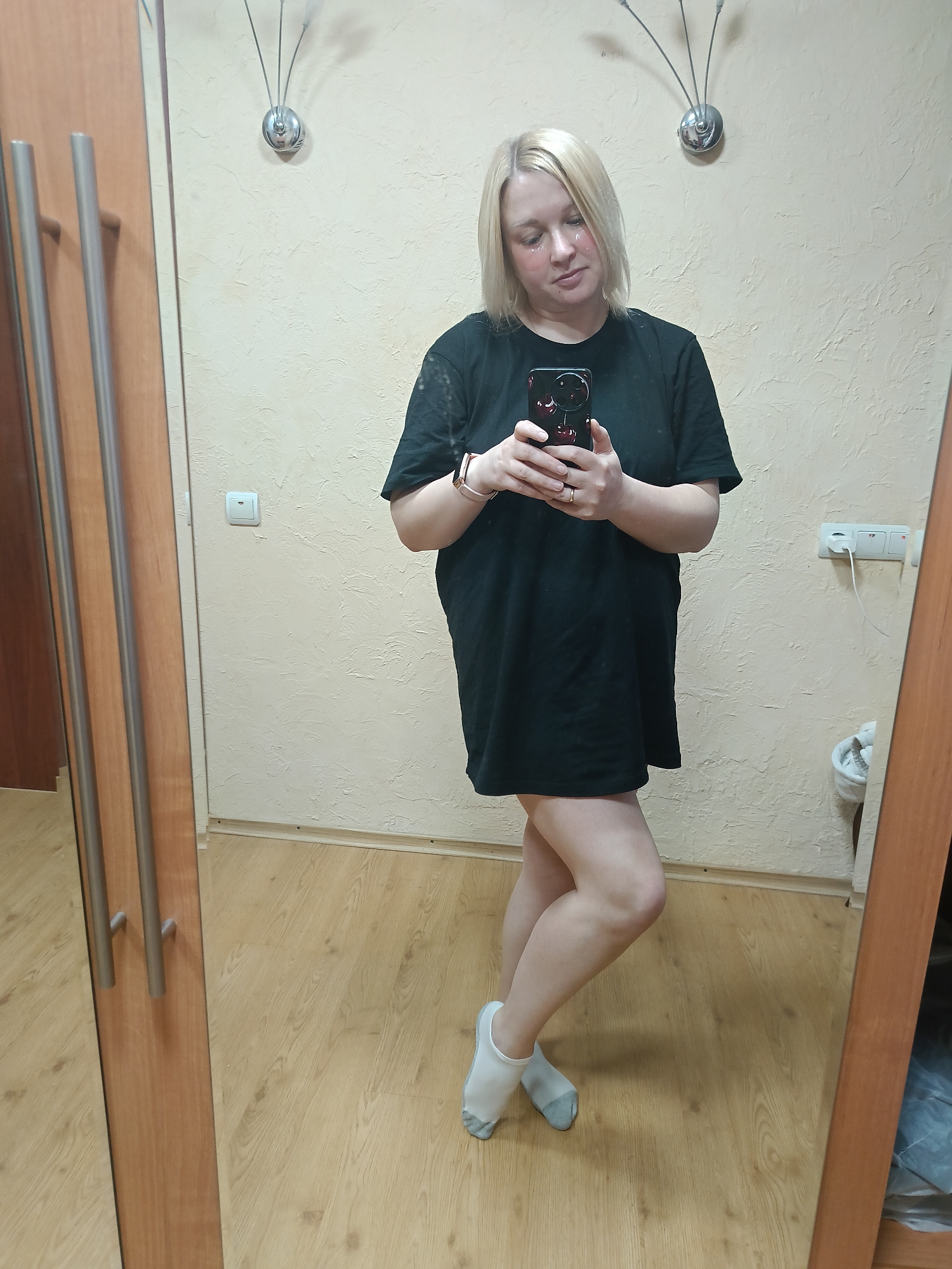 A blonde woman is depicted taking a mirror selfie in an indoor setting, likely a room or hallway in a residence. She is positioned slightly to the right of center, looking down at the black smartphone she holds in both hands, which features a distinctive red cherry design on its case. She wears a loose-fitting black t-shirt or tunic that extends to mid-thigh, revealing her bare legs, and white ankle socks with grey toes. A watch is visible on her left wrist and a ring on her right hand. Her facial expression is neutral and contemplative, with small white, possibly reflective, patches visible under her eyes, suggesting either a beauty treatment or a camera filter effect.
The background shows a light beige or cream-colored wall with a textured finish. A modern silver wall sconce with three upward-curving arms is mounted high on the wall behind her. To the left, a wooden wardrobe or closet door with two long silver handles is reflected in the mirror. To the right, a white double electrical outlet with a charging cable plugged in, and a white light switch, are visible on the wall. The floor is covered with light wooden laminate. A white cylindrical bin is partially reflected in the mirror on the far right. No discernible text is visible anywhere in the image. The scene conveys a casual, domestic moment in Chaikovsky, Russia.